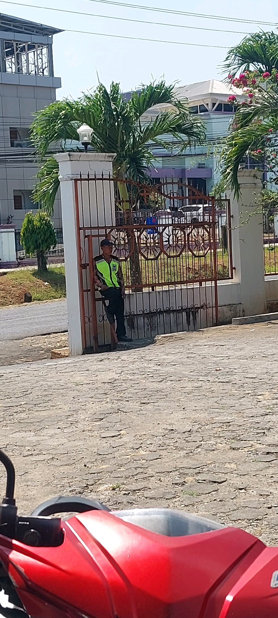 A man wearing a reflective vest stands near a decorative metal gate in Keling, Indonesia. The gate is part of a fence in front of a building. A red motorcycle is partially visible in the foreground. The scene appears to be outdoors on a paved area with some grass and trees.