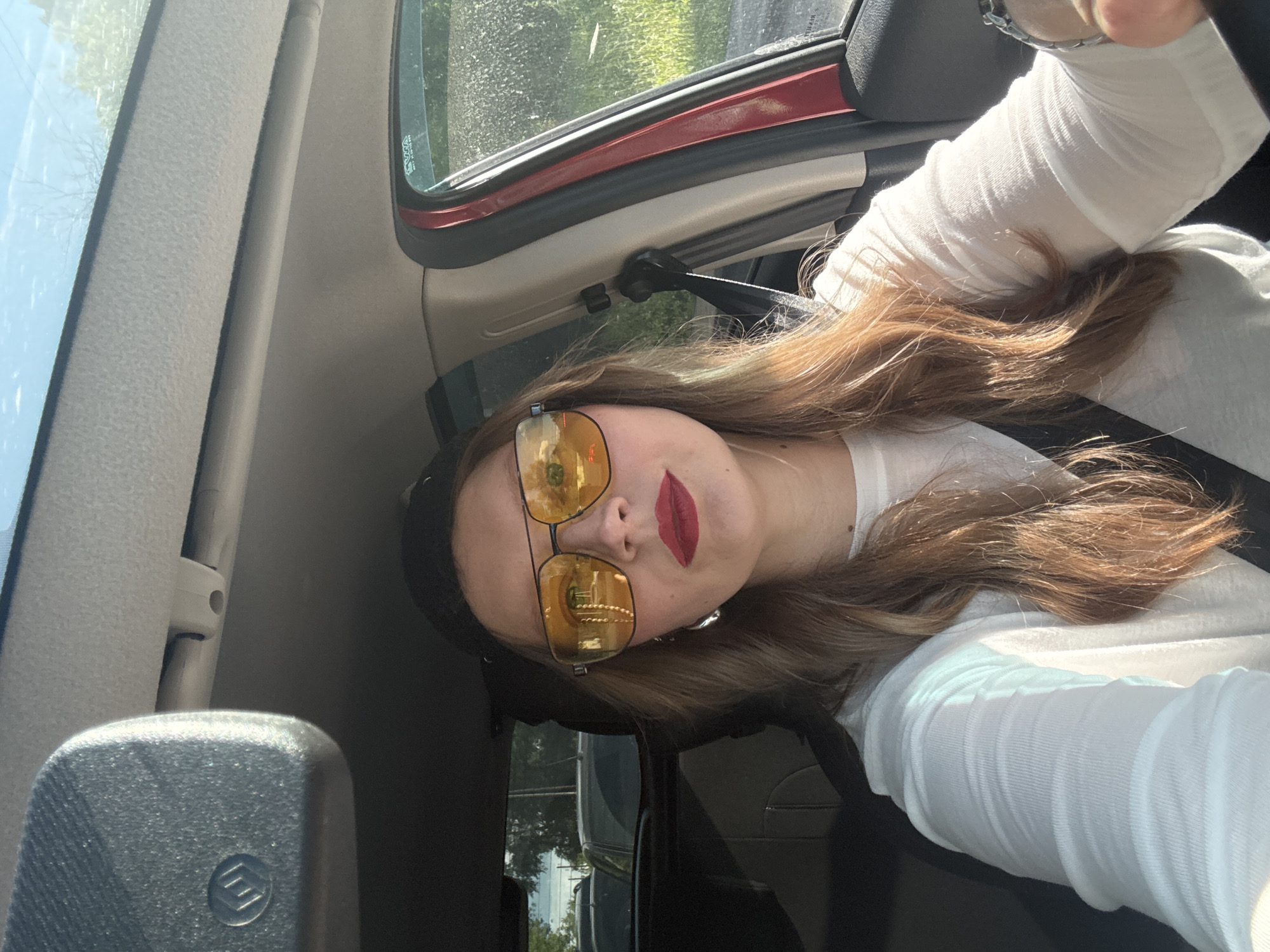 A person, likely female, takes a selfie from the driver's seat of a car, positioned as if looking up from the lower-left. The setting is bright daytime, with sunlight illuminating the car's interior. The subject has long, light brown hair flowing over their shoulder and chest. They are wearing a white long-sleeved top and a dark headband or hat.
Their face is partially visible, featuring prominent red lipstick. Large, gold-framed aviator-style sunglasses with yellow-tinted lenses cover their eyes, reflecting green foliage and parts of an unseen structure from outside. The car's interior shows a gray or beige A-pillar and headliner, with a red trim accenting the side window frame. A seatbelt mechanism is visible near the red trim, and a portion of the steering wheel can be seen on the upper right.
Through the car windows, green trees and blue sky are visible, suggesting an outdoor, possibly suburban or natural, environment. A small, circular black logo, possibly stylized as an "M" or three horizontal lines within a circle, is present on a black plastic component in the lower-left foreground, likely part of the rearview mirror housing. Faint white text, including "VWA" and "DOT," is visible on a sticker or etching on the car's upper right window. The car appears to be a left-hand drive vehicle. The image captures a casual moment in Izhevsk, Russia.