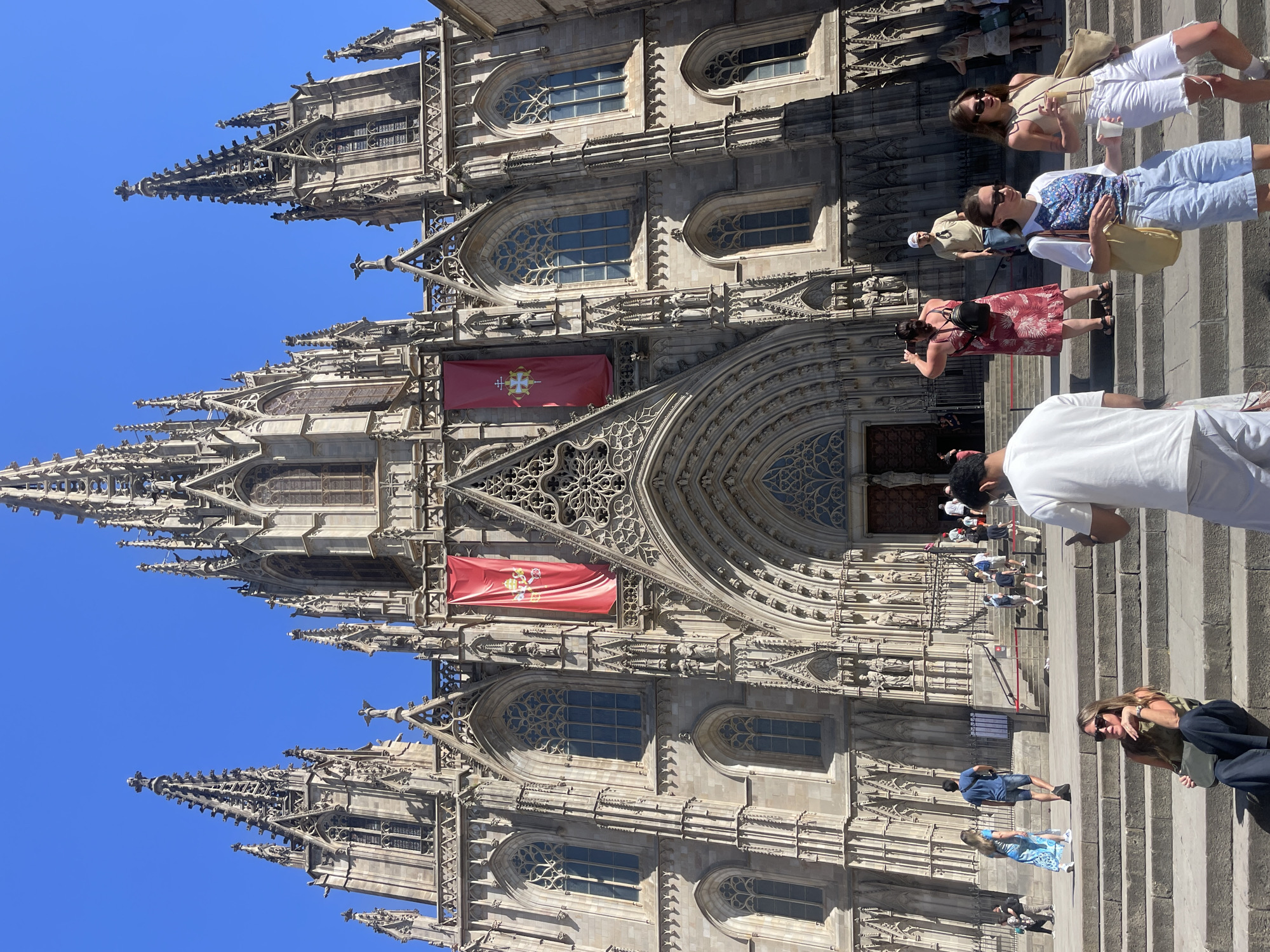 The image depicts the Barcelona Cathedral, a Roman Catholic cathedral dedicated to the Holy Cross and Saint Eulalia, in Barcelona, Spain. The cathedral's Gothic facade is visible, with intricate architectural details and spires reaching towards a clear blue sky. Two large red banners with a golden emblem are hung on either side of the main entrance. A crowd of tourists are gathered on the steps leading up to the cathedral. Several individuals are seated on the steps, while others are walking up or standing. The scene is bathed in sunlight, indicating a warm, clear day.