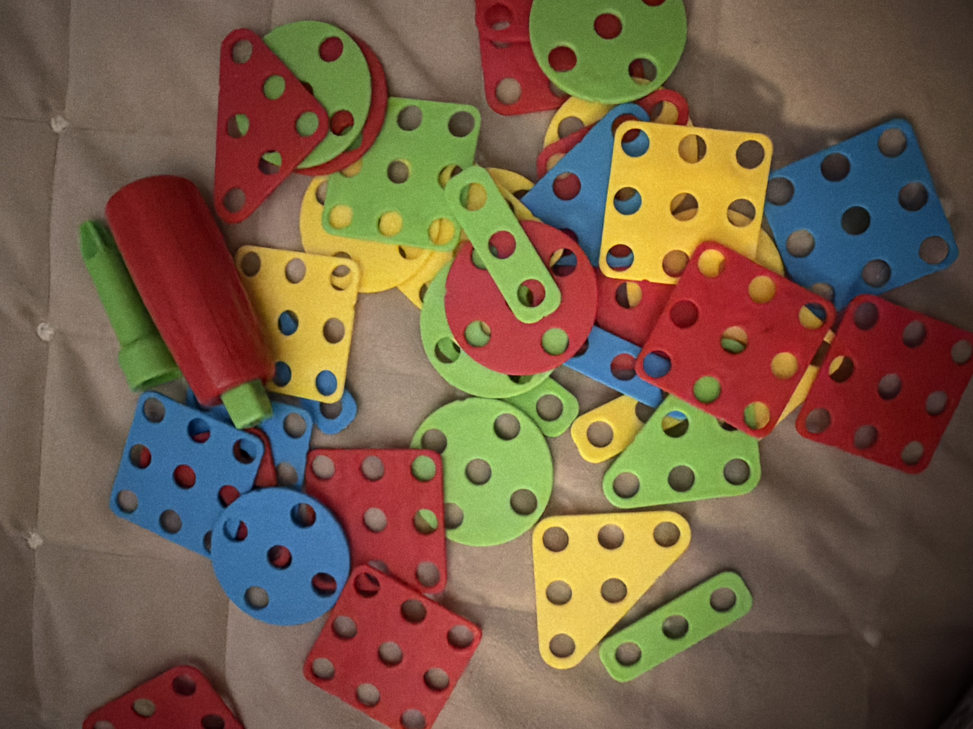 The image shows a collection of children's construction toys scattered on a light brown fabric surface. The toys are made of colorful foam, primarily in red, blue, yellow, and green. The shapes include squares, circles, triangles, and rectangular bars, all with circular holes. There also is a red and green screwdriver toy. The lighting is somewhat subdued, creating shadows on the fabric. There is no evidence of people, actions, or indications of a specific location such as Votkinsk, Russia.