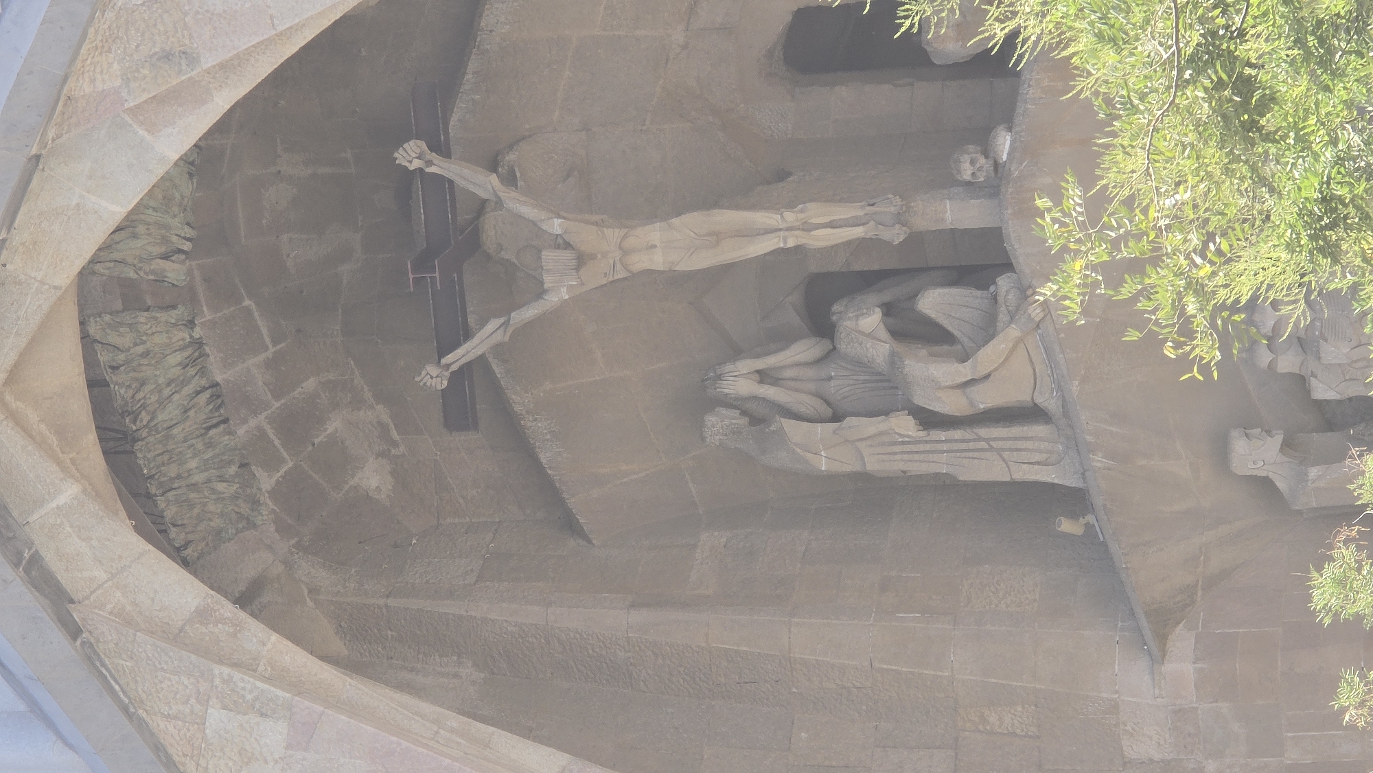 The image shows a detailed stone sculpture of the crucifixion of Jesus Christ, located on the facade of the Sagrada Familia in Barcelona, Spain. Jesus is depicted on a cross, with his arms outstretched and body emaciated. Below the cross, several figures are shown in various poses of grief and lamentation. One figure, presumably Mary, is shown with her hands covering her face, while another figure kneels. The sculpture is carved into the stone facade of the basilica, which features intricate architectural details and arched openings. The overall tone of the sculpture is somber and emotional, conveying the pain and sorrow associated with the crucifixion. The image is captured from a slightly low angle, emphasizing the scale and grandeur of the basilica's facade. Green foliage is visible in the foreground, partially obscuring the lower portion of the sculpture.