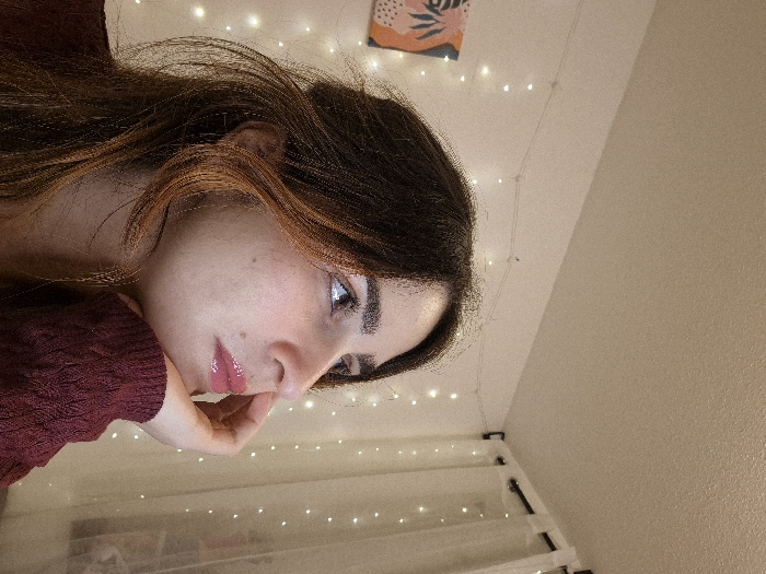 A young woman with dark hair and fair skin rests her chin on her hand, gazing thoughtfully to the side. She wears a dark red, textured top. Her lips appear glossy, and her features are soft. Behind her, string lights illuminate a white surface, possibly a headboard, with another set of lights strung along the wall. A small art print adorns the upper left corner of the scene, which has been rotated slightly. The background features a plain, light-colored wall.