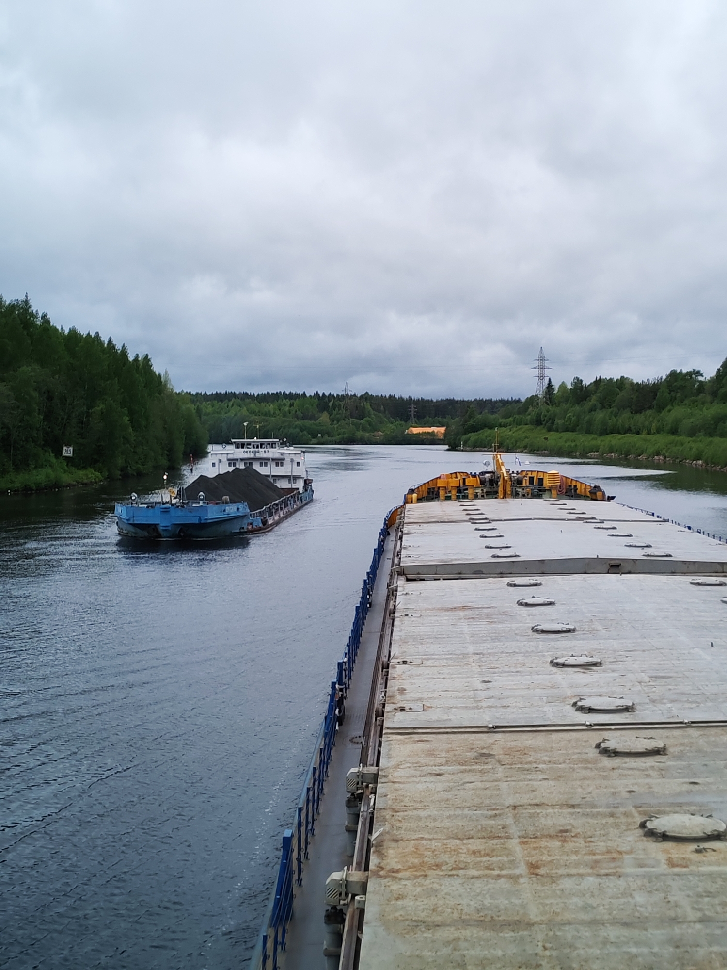 The image shows a wide canal with two barges. One barge, closer to the viewer, has a flat, gray deck with numerous circular hatches. The second barge, further away, is loaded with a dark substance, possibly coal, and has a blue hull and a white cabin. Both barges are moving through the water, leaving wakes behind them.
The canal is lined with lush green trees on both sides. In the distance, there is a transmission tower visible against an overcast sky.
Based on the location, it is likely that this scene depicts the Volga-Baltic Waterway in Russia, near the city of Depo.