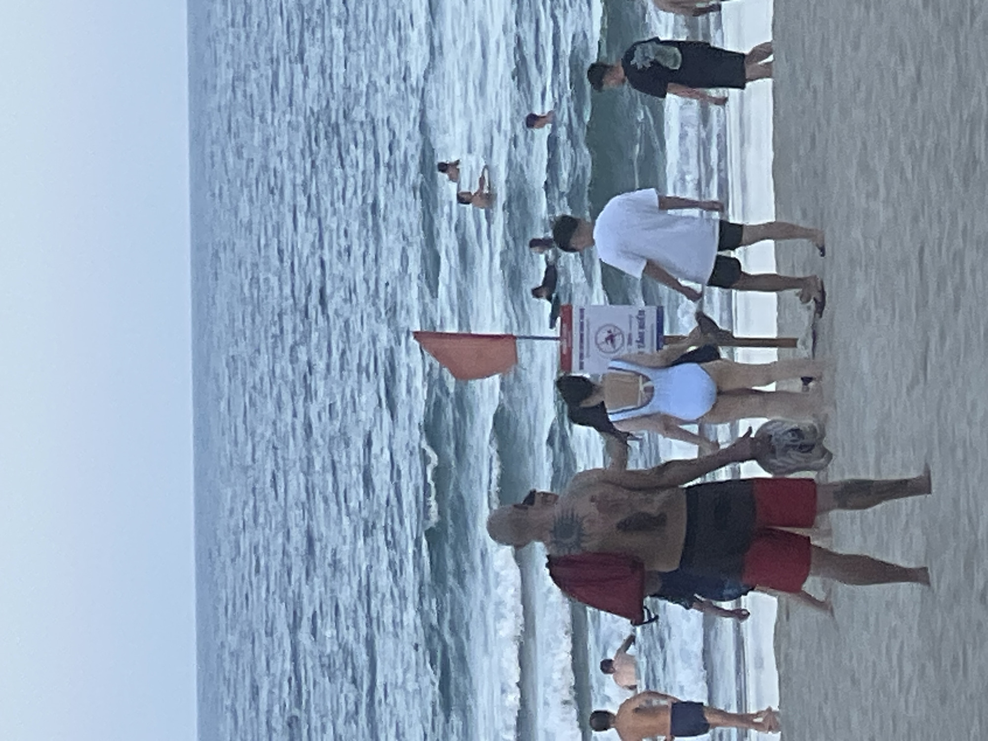 The image depicts a beach scene in Đà Nẵng, Vietnam, likely during the late afternoon or early evening. The ocean is calm with moderate waves, and the sky is overcast. Several people are visible in the water, some swimming and others with surfboards. On the beach, a group of individuals are gathered, some walking and others standing. In the foreground, a man with extensive tattoos on his back is seen from behind, wearing red and black shorts and carrying a red bag over his shoulder. To his right, a woman in a light blue swimsuit stands facing away from the camera. A red flag on a pole, along with a sign, is also visible on the beach, indicating a potential hazard or a designated swimming area. The overall atmosphere appears relaxed, with people enjoying a day at the beach.