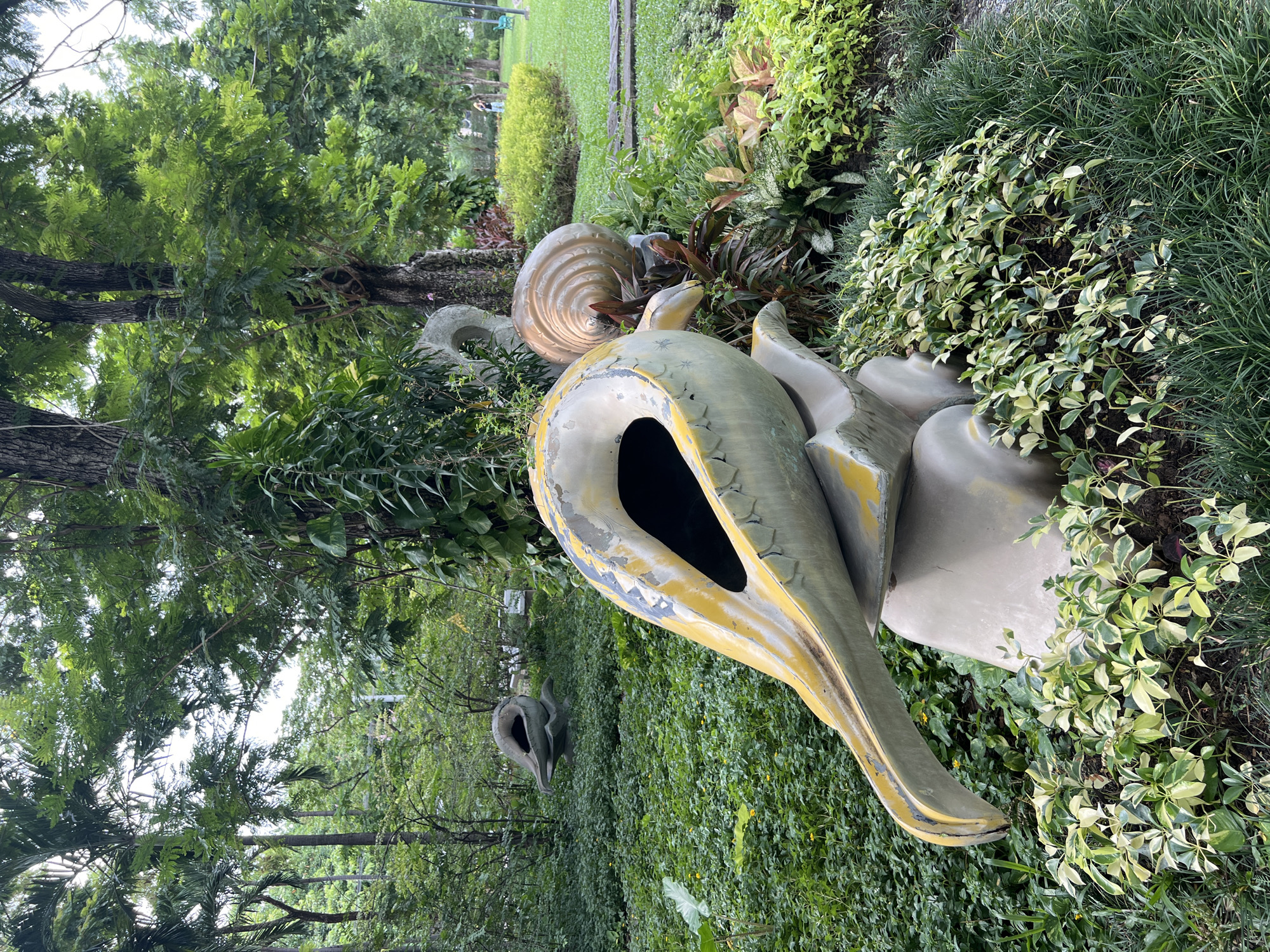 In a verdant park setting in Bangkok, Thailand, large abstract sculptures resembling shells are situated amidst lush greenery. The foreground features a prominent sculpture, predominantly light grey with yellow accents, shaped like a curled shell with an open cavity. It rests on a similarly colored base. Behind it, a larger, more detailed sculpture of a coiled shell, golden-brown in color, is visible. Further in the background, another shell-like sculpture is nestled in the dense foliage. The environment is characterized by mature trees with dense canopies, providing shade over a carpet of various plants and grasses. Sunlight filters through the leaves, creating dappled light on the ground. In the distance, a paved path winds through the park, and a few individuals can be seen walking, one of whom is a person in a light-colored top and dark bottoms. The overall atmosphere is serene and natural, with the sculptures integrated as artistic elements within the landscape.