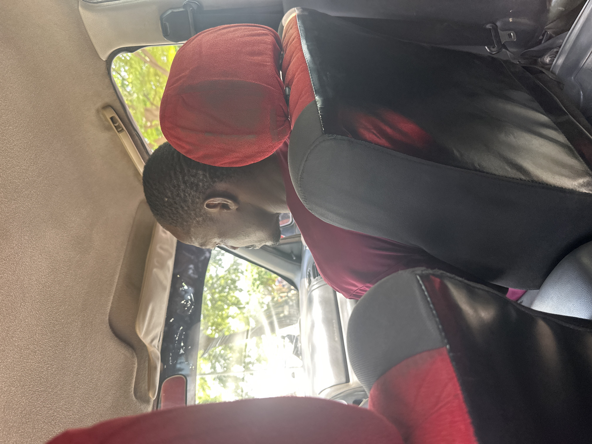 The image shows the interior of a vehicle, likely a car or van, seen from a low angle. The focus is on a young person with short hair and dark skin, wearing a dark red shirt, who is leaning their head against a red headrest. They appear to be resting or looking down. The headrest is plush and textured.
The seats are a mix of black and red material, with some grey trim. The interior also shows the fabric lining of the car's roof and pillar, which is a light beige color. Through the window, which is partially visible and reflecting light, a bright, sunny daytime scene of lush green foliage can be seen. The sunlight is strong, creating a glare on the window and obscuring some details outside.
The setting appears to be urban or suburban, given the presence of a vehicle's interior. The bright daylight suggests it is daytime, with clear weather. The image captures a moment of stillness within the moving environment of a vehicle. There is no visible text in the image. The overall mood is casual and everyday. The location context is Abuja, Nigeria.