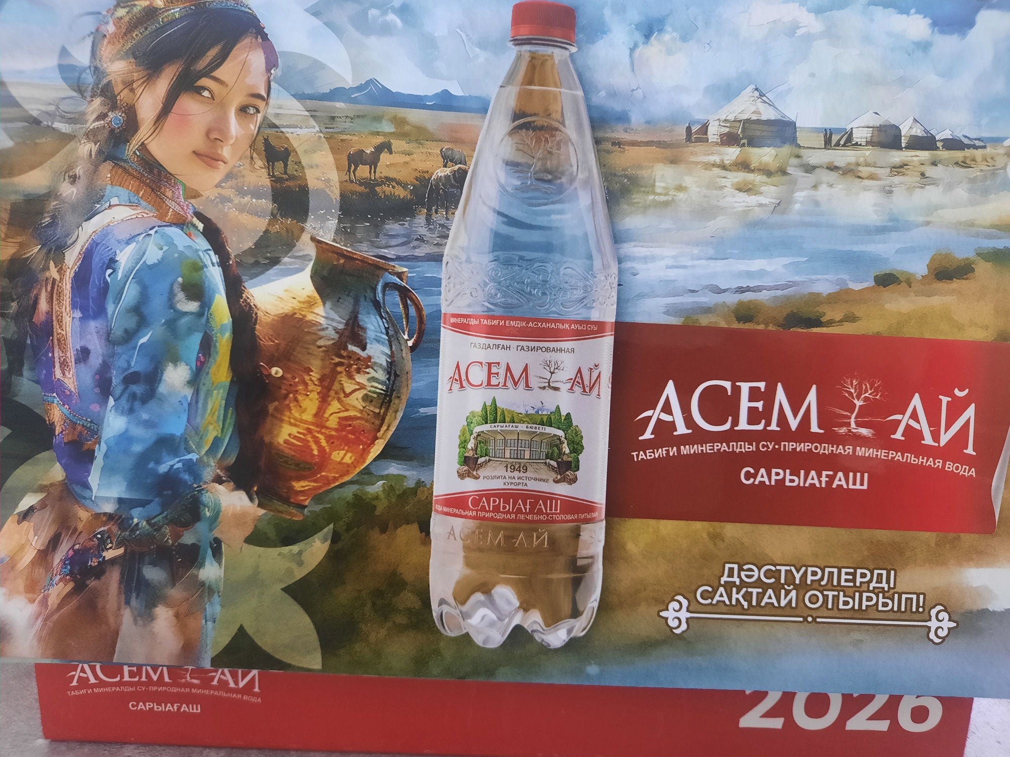 This is a promotional image for "ACEM-AI" mineral water, featuring a woman in traditional clothing on the left and a bottle of water prominently displayed. The background depicts a pastoral landscape with yurts, horses, and mountains, evoking a sense of nomadic heritage. The text on the image is in Kazakh and Russian. A portion of the image shows "2026", which is likely the year the advertisement is intended for. While the advertisement itself does not specify a location in Zhanaozen, Kazakhstan, it is likely being displayed or distributed within the city as part of a marketing campaign.