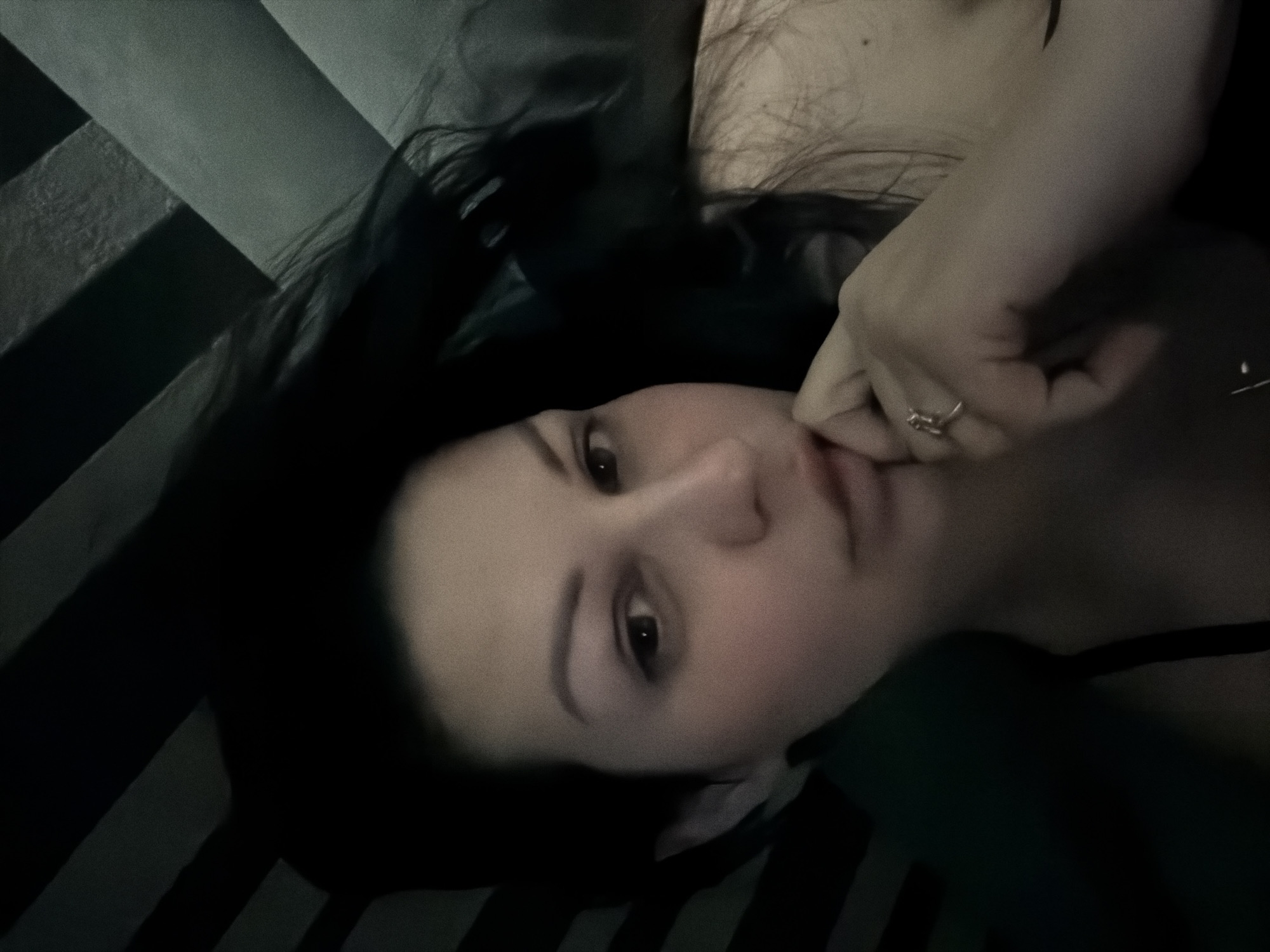 A low-light, intimate portrait captures a young woman lying down in an indoor setting, likely in Sochi, Russia. Her dark, long hair is spread around her head, contrasting with her fair skin. She has dark, expressive eyes that gaze slightly to the right of the camera. Her right hand is raised near her chin or mouth, with her fingers gently touching her face, and a subtle ring is visible on her ring finger.
The woman is positioned against a background of dark and lighter stripes, possibly a bedspread, pillow, or upholstered surface. The dim lighting creates soft shadows and a subdued, somewhat pensive mood. There are no clear indications of the time of day, but the low light suggests it could be evening or night. No specific weather details are discernible. The woman's expression is neutral, bordering on contemplative or serene. There is no visible text in the image.