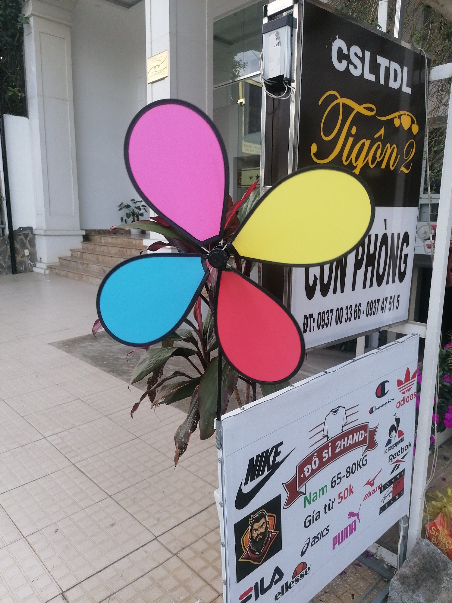 The image captures a colorful, four-petal pinwheel standing in front of a store sign in Da Lat, Vietnam. The petals of the pinwheel are distinctively colored pink, yellow, blue, and red, each with a black outline. Behind the pinwheel is a sign with the words "CSLTDL Tigon 2" in large, stylized text above "CON PHONG". A phone number is printed below.
In the foreground, a smaller sign displays logos of various apparel brands such as Nike, Champion, Adidas, Reebok, Puma, Fila, and Ellesse, along with the words "ĐỒ SĨ 2HAND" and "Nam 65-80KG Giá từ 50k". The tiles of the pavement are a pale tan color. The surroundings include building columns, stairs leading to a glass door, and some plants.