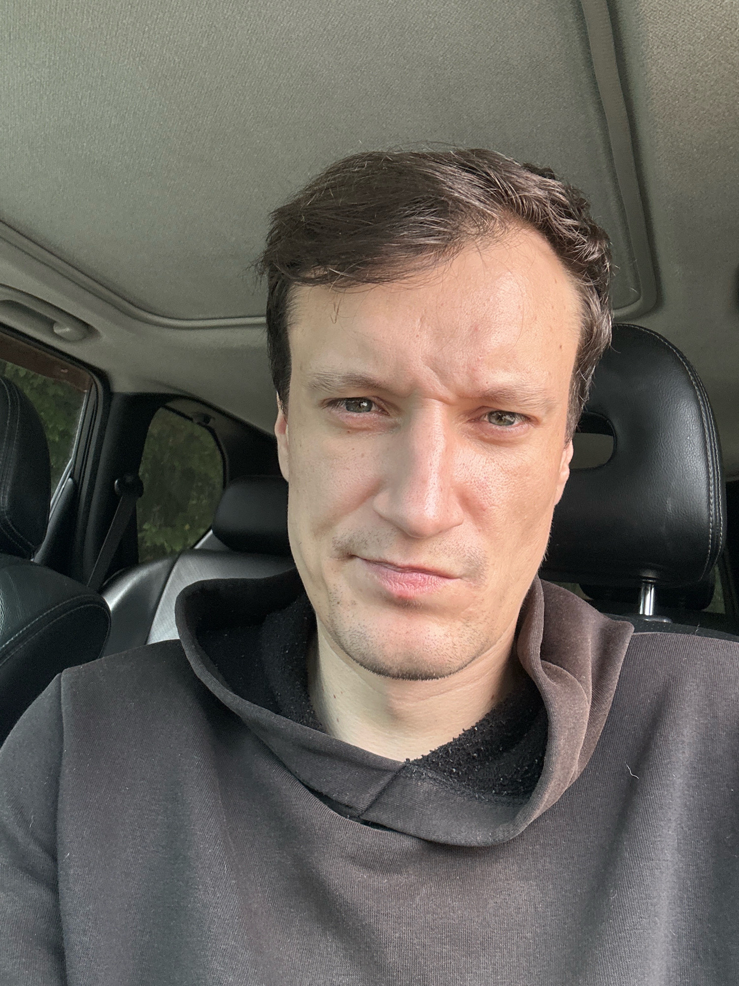 A selfie taken from a high angle shows a man with light brown hair, fair skin, and green eyes. He is wearing a dark brown hoodie and appears to be in the driver's seat of a car. The car's sunroof is open, and foliage can be seen through the rear window, suggesting an outdoor setting. The man's facial expression is ambiguous, with his eyebrows slightly furrowed and his mouth in a neutral or slightly downturned position. The background includes the car's interior, specifically the headrest of the passenger seat and part of the dashboard. The lighting is natural, indicating daylight. The image does not contain any explicit geographical markers or clues to pinpoint its location within the city of Korchivka, Ukraine.