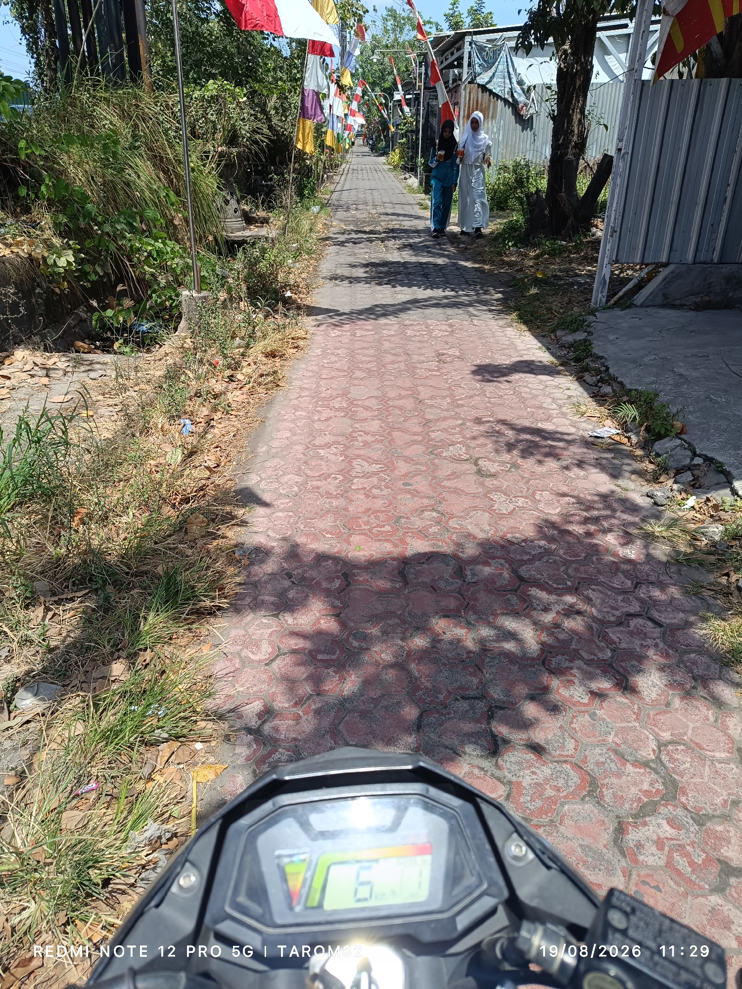 This image depicts a sunlit street in Tebel, Indonesia, likely on August 19, 2026, at 11:29 AM, as indicated by the timestamp overlay. The perspective is from a motorcycle, with its dashboard visible in the foreground, displaying a speed of 6 km/h. A paved path, made of red hexagonal bricks, stretches into the distance, lined on the left by dry grass, fallen leaves, and dense green vegetation. On the right side of the path, corrugated metal fencing and trees are visible. Two individuals, possibly women, are walking away from the camera on the path. They are dressed in modest attire, one in a blue tunic and pants, and the other in a white headscarf and a long white dress. Flags, red and white, are strung along poles, suggesting a local celebration or event. The overall environment is outdoor, with clear daylight casting shadows across the path.