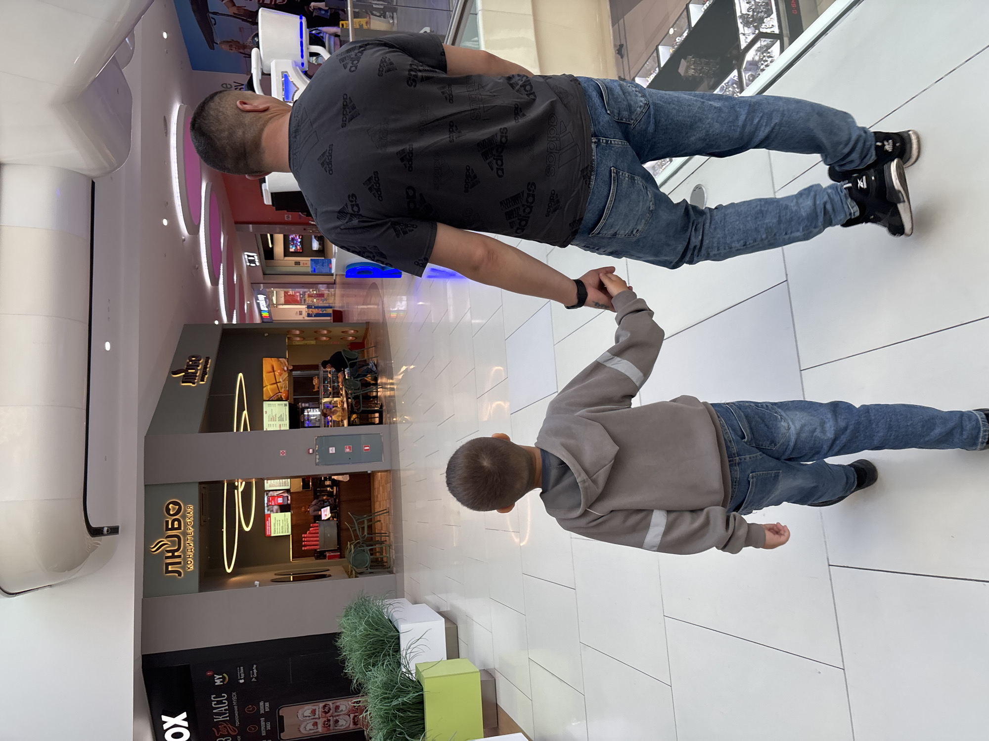 In a brightly lit indoor shopping mall, a man and a young boy walk hand-in-hand. The man, seen from behind, wears a dark grey t-shirt with a repeated Adidas logo pattern and light blue distressed jeans. He has a black smartwatch on his left wrist. The boy, also viewed from behind, wears a light grey hooded sweatshirt with reflective silver stripes on the sleeves and blue jeans. They are walking on a polished white tiled floor. To their left, a storefront with a prominent "ЛЮБО" (Lyubo) logo is visible, appearing to be a cafe or confectionery. The environment suggests a modern commercial space with large windows and overhead lighting. The location is identified as Tlustenkhabl, Russia.