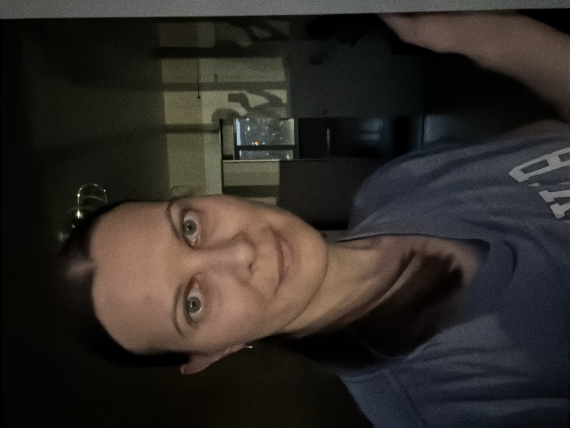 A woman with dark hair pulled back and a light blue t-shirt takes a selfie in what appears to be an apartment or hotel room. She is looking directly at the camera with a neutral expression. The background shows a window with a cityscape visible, suggesting the location is an urban area at night. Shadows of the woman and the person holding the camera are cast on a reflective surface. The overall lighting is dim, with some artificial light sources from the city outside. The woman is wearing a small hoop earring. The image quality is grainy.