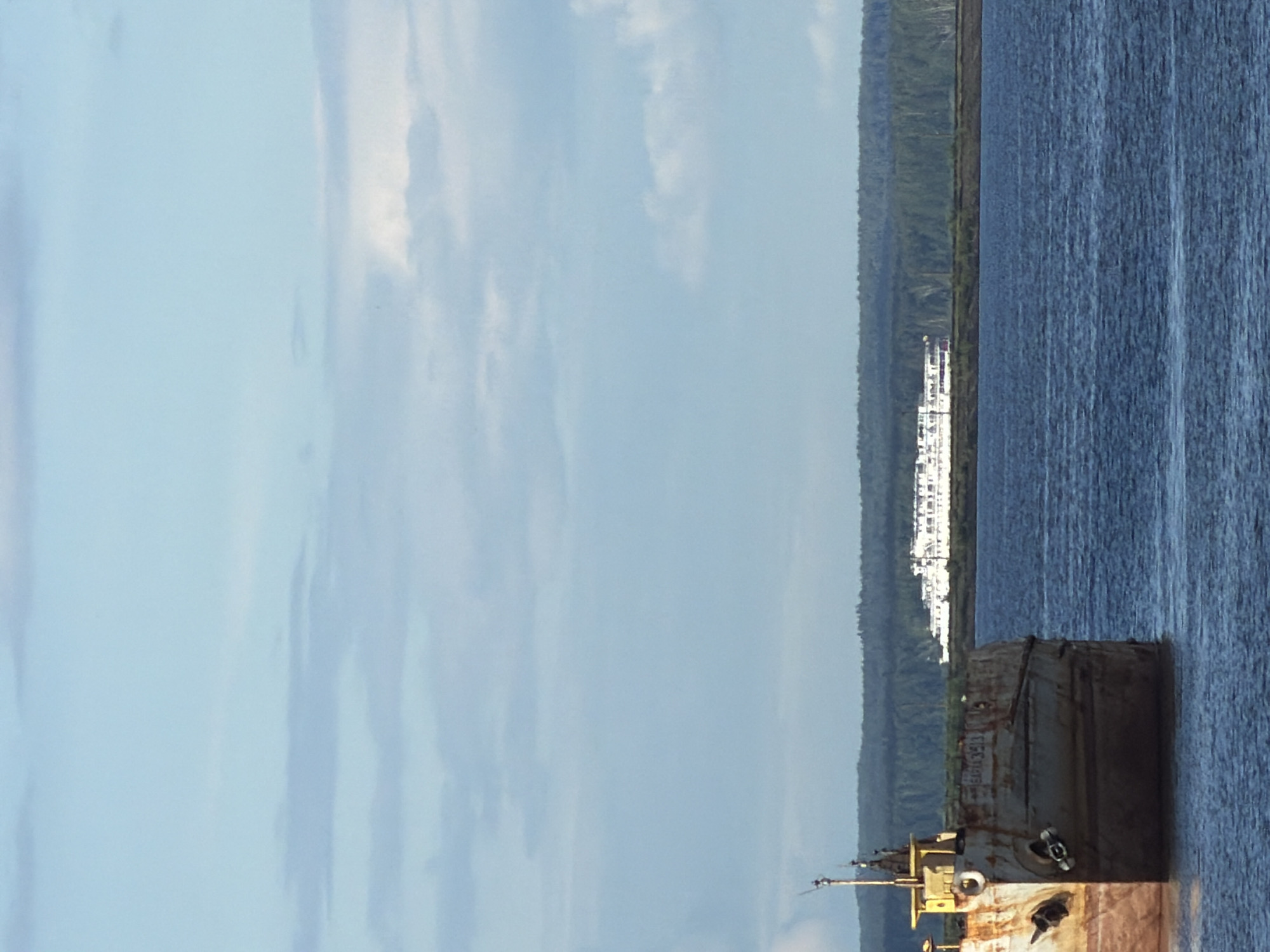 The image depicts a river scene in Chaikovsky, Russia. A large, dilapidated barge occupies the lower right quadrant of the frame, partially submerged in the water. Beyond the barge, a body of water extends towards the middle of the image, with a dense, green forest lining the opposite shore. Further in the distance, a cluster of white buildings is visible against the backdrop of the forest. The sky, which takes up the majority of the image, is pale blue with streaks of white clouds.