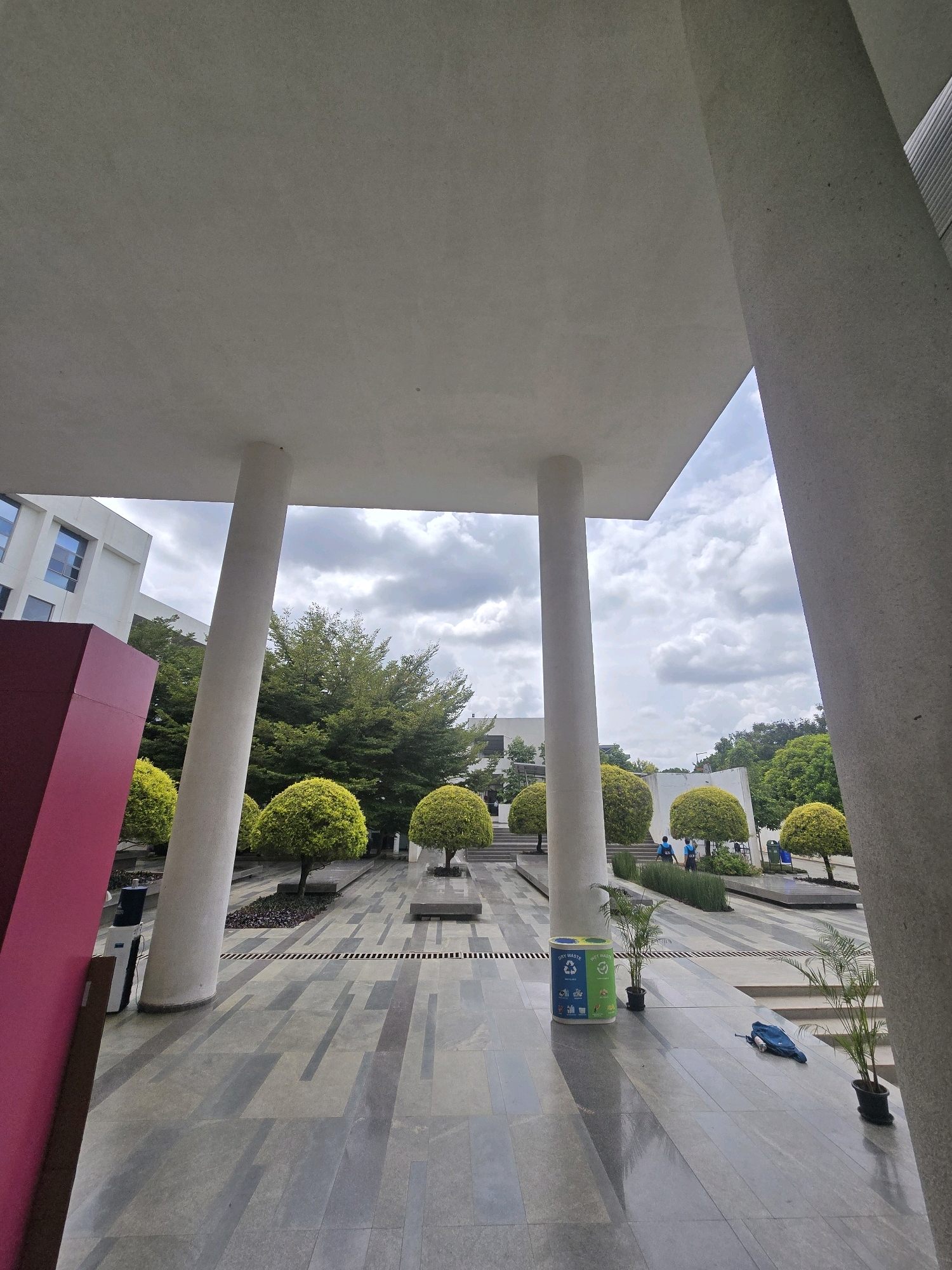 The image depicts an outdoor courtyard with modern architecture and landscaping. In the foreground, a large, light-colored concrete structure, possibly a canopy or building overhang, casts shadows across a paved ground area. The ground is made of large, rectangular stone tiles in varying shades of gray and dark gray, arranged in a geometric pattern. Several thick, white, cylindrical columns support the overhead structure, one of which is prominently positioned in the center. To the left, a large, angled, magenta-colored rectangular object protrudes into the frame.
Beyond the immediate foreground, the courtyard opens up to a landscaped area with several spherical, topiary trees with vibrant green foliage. These trees are planted in raised beds or on platforms. In the background, a modern building with white walls and several horizontal, rectangular windows can be seen. Lush green trees form a backdrop behind the manicured bushes.
Two individuals are visible in the middle ground, appearing to be walking or standing near the trees. One is wearing a blue shirt and the other a dark blue shirt. A blue and green recycling bin with compartments labeled "DRY WASTE" and "WET WASTE" is situated on the pavement near one of the columns. A dark blue bag or piece of luggage lies on the ground to the right of the recycling bin, near a potted plant. The sky above is partly cloudy with a mix of gray and white clouds. The overall impression is a well-maintained, contemporary urban outdoor space.
The image is taken at the Infosys Foundation, located in Ittagalapura, India.