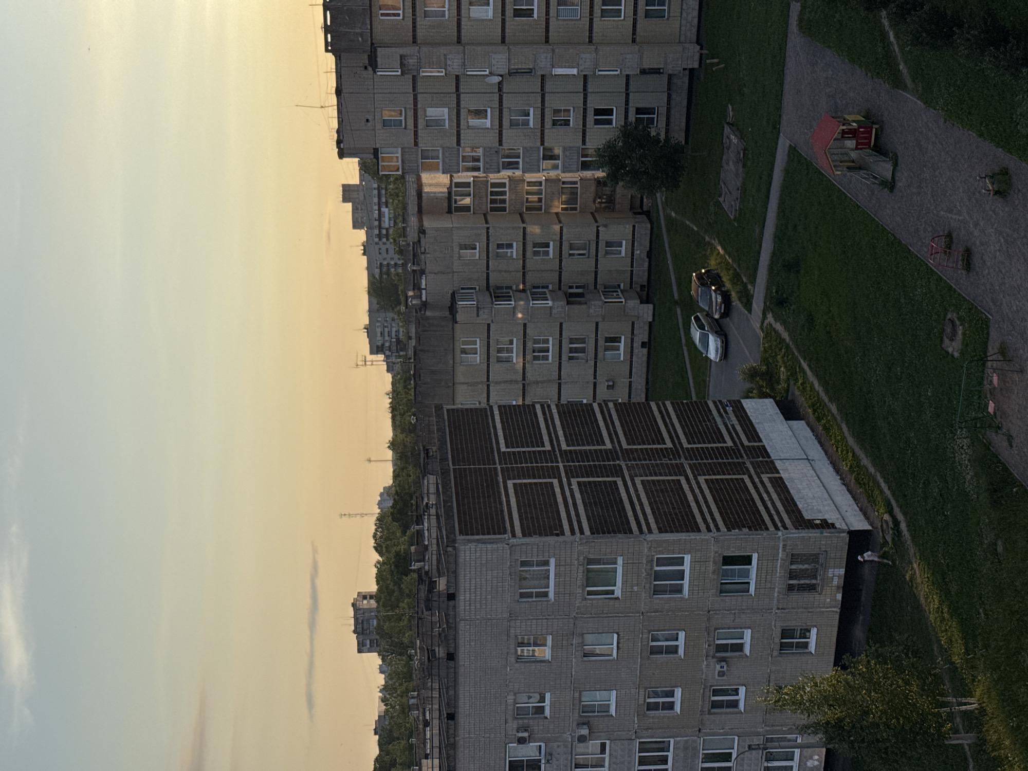 The image captures a twilight view of a residential area in Kirishi, Russia. The sky is a soft gradient of pale blue and warm orange, indicating the setting sun. In the foreground, two multi-story apartment buildings, constructed with grey brick and featuring numerous windows and balconies, dominate the scene. The building on the left has a distinct geometric pattern of dark brown panels on its facade. In front of the buildings, there is a patch of green grass, a paved walkway, and a small children's play area with a red and yellow playhouse. Two cars, a silver sedan and a dark SUV, are parked on the asphalt. A lone figure in light-colored clothing is walking on the grass pathway. In the background, more apartment buildings and trees are visible, with utility poles and distant structures also discernible. The overall impression is a quiet, ordinary evening in a Soviet-era housing complex.