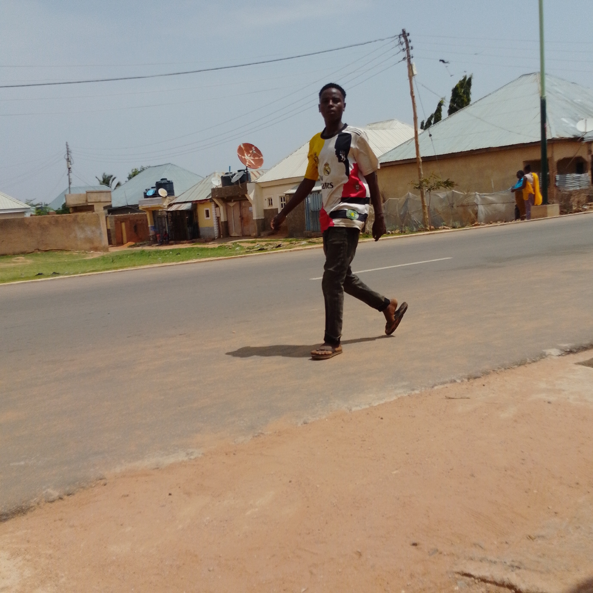 In Gombe, Nigeria, a young Black man is captured mid-stride on a paved road with a dirt shoulder. He is wearing a white, yellow, black, and red t-shirt with intricate designs and dark pants, along with brown sandals. His shadow is cast to his left on the road. Behind him, the environment consists of various residential buildings with corrugated metal roofs, one of which features a satellite dish. In the distance, two other individuals are visible near a building. The sky above is clear and bright, suggesting a sunny day.