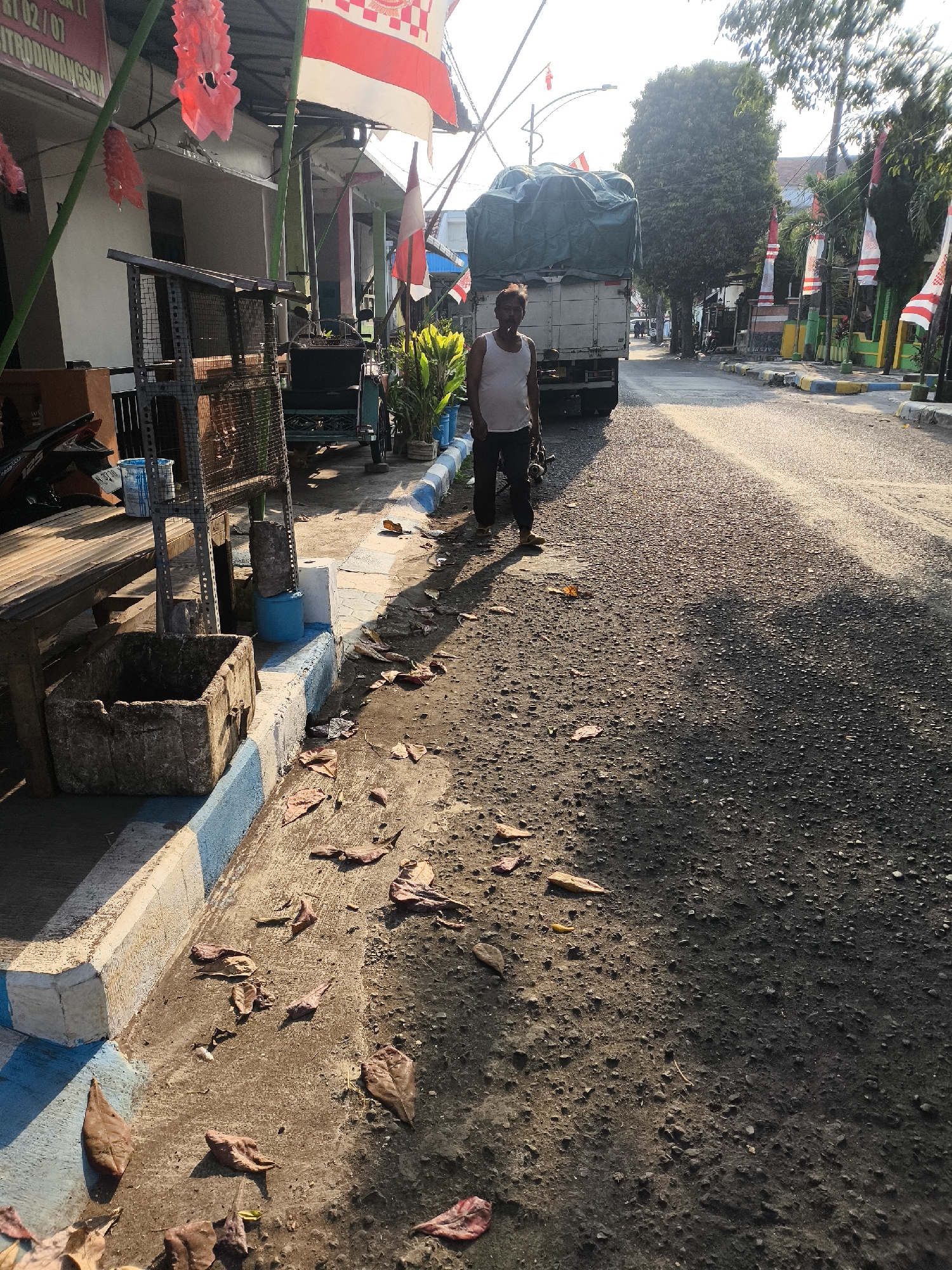 The image depicts a street scene in the city of Citrodiwangsan, Indonesia. In the foreground, a man in a white tank top and dark pants stands on the side of a paved road. The road is textured with small stones and appears to be partially unpaved in some areas. Dry leaves are scattered on the ground, particularly along the edge of the road and sidewalk.

To the left of the man, a large truck with a green tarp covering its cargo is parked. A bench made of wood is visible next to a metal rack structure. A concrete planter box is positioned near the bench. Blue and white painted curbs line the edge of the sidewalk.

In the background, buildings and trees line the street. Several flags, including Indonesian flags and decorative red and white banners, are displayed along the roadside. A motorcycle is parked near the buildings. The overall lighting suggests it is daytime, possibly late afternoon, with shadows cast across the scene. The environment appears to be a somewhat commercial or residential area within the city.