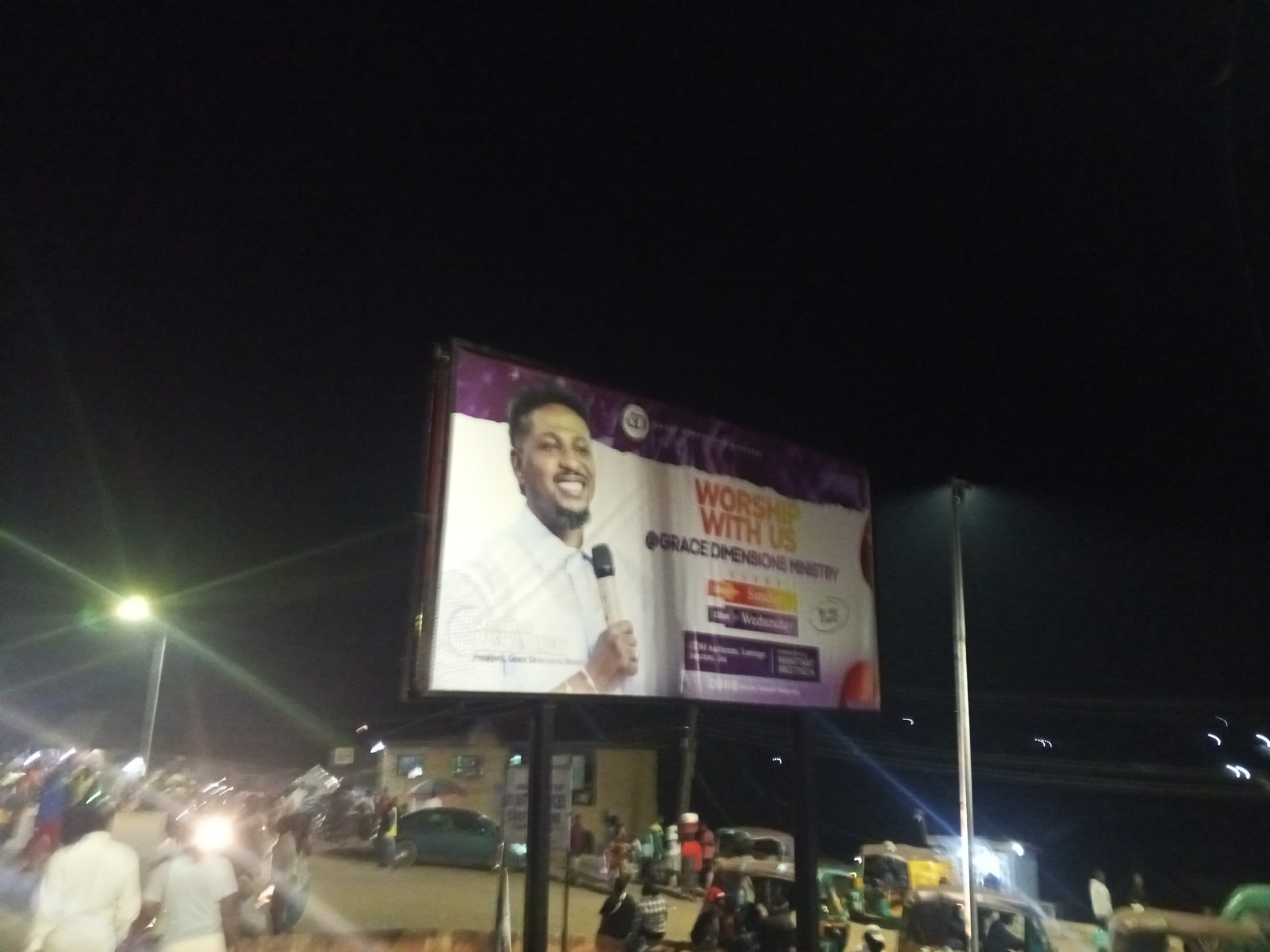 A photograph taken at night depicts a street scene in Gwong, Nigeria. Dominating the view is a large billboard advertising "Worship With Us" at Grace Dimensions Ministry, featuring a smiling man holding a microphone. The billboard lists service days as Sundays and Wednesdays. Below the billboard, a street teeming with people and vehicles is visible. The people appear to be walking and standing around. The vehicles include cars and what appear to be auto-rickshaws. Streetlights illuminate the scene, casting a glow on the road and surrounding buildings. The lighting conditions suggest it is late in the evening or early night.