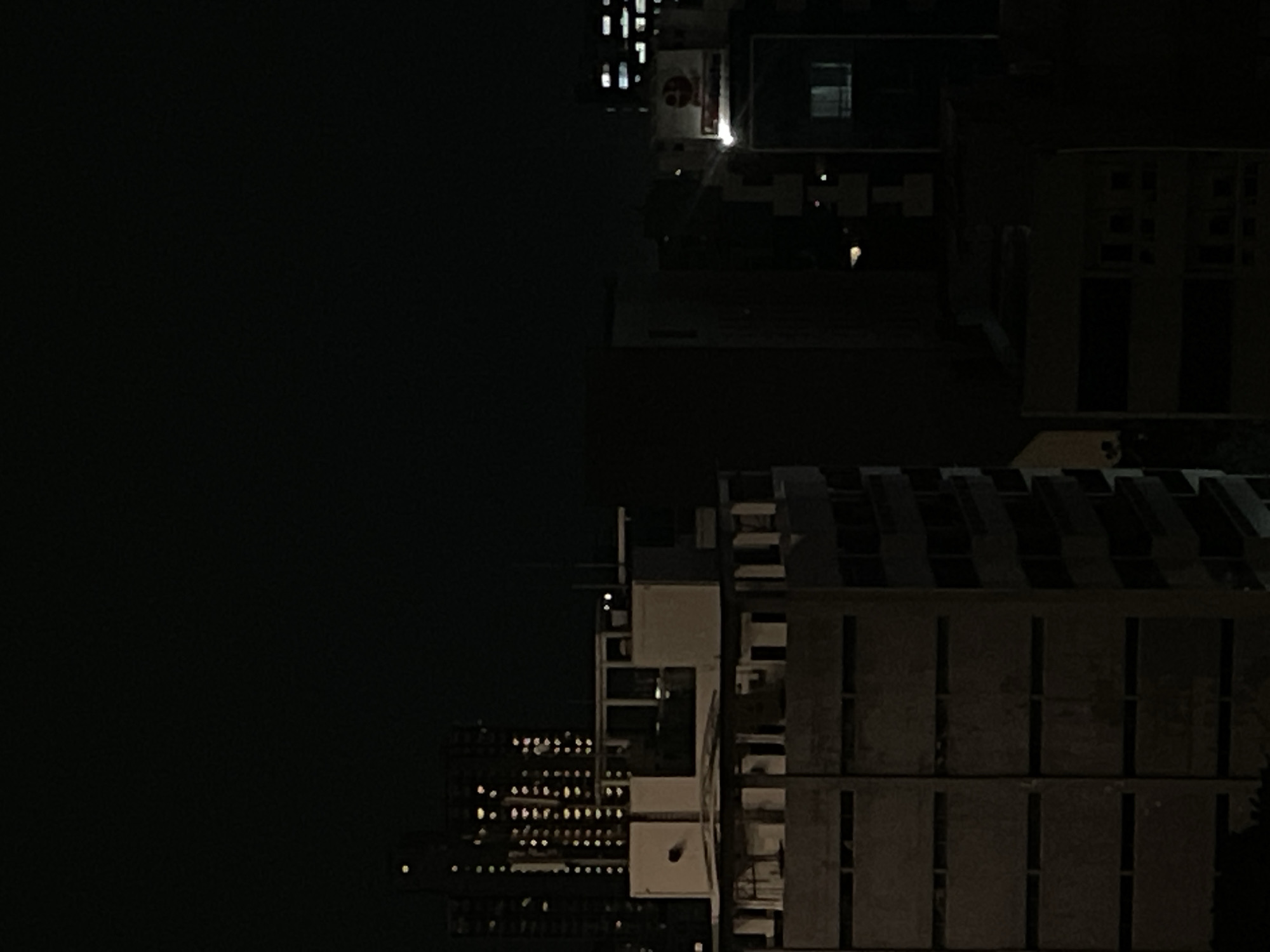 The image depicts an aerial view of buildings in Central Jakarta, Indonesia, at night. The left portion of the image is dominated by darkness, likely representing the night sky. The right portion shows a cluster of buildings with lit windows. The buildings vary in height and structure. Some buildings have rows of windows while others show limited lighting.
