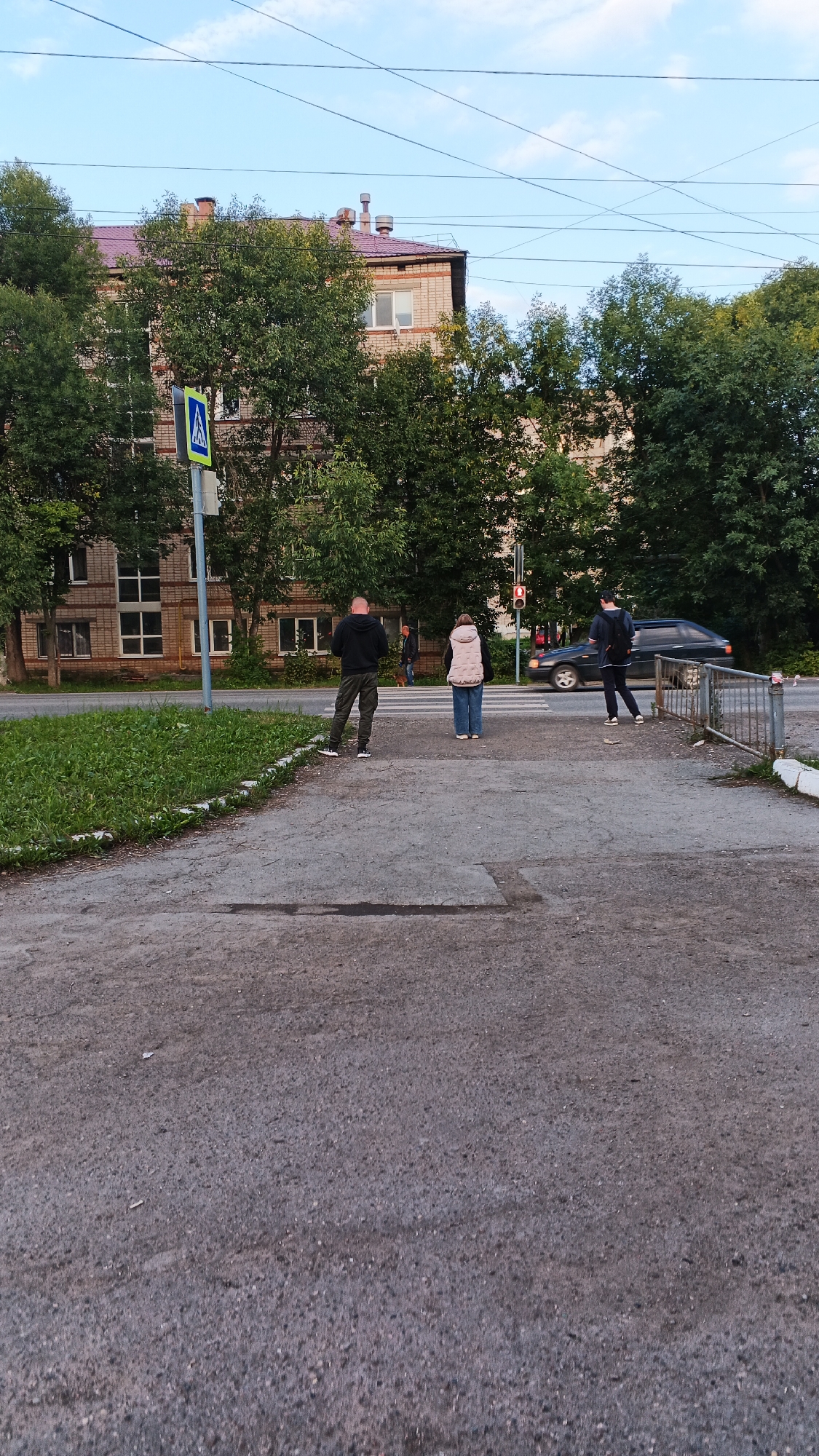 In Izhevsk, Russia, a sidewalk and pedestrian crossing area is depicted under a partly cloudy sky. The scene includes a brick apartment building with a reddish-brown roof and several trees. A pedestrian crossing sign is visible on the left side of the image. Three individuals are present: one man, a woman, and another person wearing a backpack. A dark-colored car is driving across the pedestrian crossing. A metal fence is also visible on the right side of the image. The ground is a mixture of asphalt and concrete.