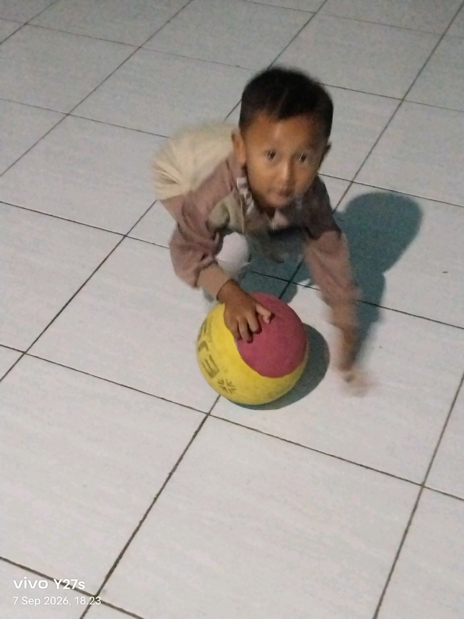 An overhead medium shot captures a young boy on his hands and knees on a tiled floor, his body angled towards the bottom right of the frame. He is wearing a long-sleeved, light brown shirt with white accents and light-colored shorts. His face, turned towards the camera, shows a clear gaze and his short, dark hair is neatly cut. A red and yellow ball rests to his right, partially obscured by his body. The floor is covered in large, square, light-colored tiles with dark grout lines, creating a grid pattern. A shadow of the boy and the ball is cast on the floor to the right. The bottom left of the image shows a timestamp and device information: "vivo Y27s 7 Sep 2026, 18.23". The scene appears to be indoors, possibly a home or a community space in the city of Tamansari, Indonesia.