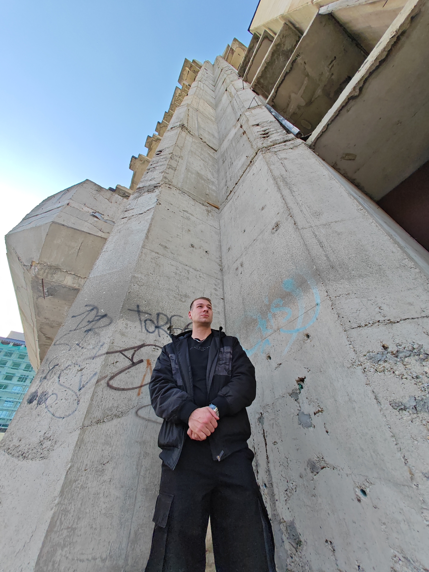 A male individual stands in front of a massive, unfinished or dilapidated concrete structure in Shumen, Bulgaria. The structure, constructed from raw grey concrete, features angular projections, exposed rebar, and signs of weathering and incomplete construction. Graffiti in black and light blue spray paint is visible on the concrete surfaces. The subject, appearing to be a young adult, is dressed in a black bomber jacket with subtle patterned sleeves, a black t-shirt, and black cargo pants. A silver-colored wristwatch is on his left wrist. He is positioned facing slightly left, looking upwards and to his left, with his hands clasped together in front of him. The photograph is taken from a low angle, emphasizing the towering height of the concrete edifice against a clear blue sky. In the distant left background, a modern building with green-tinted windows and scaffolding is partially visible.