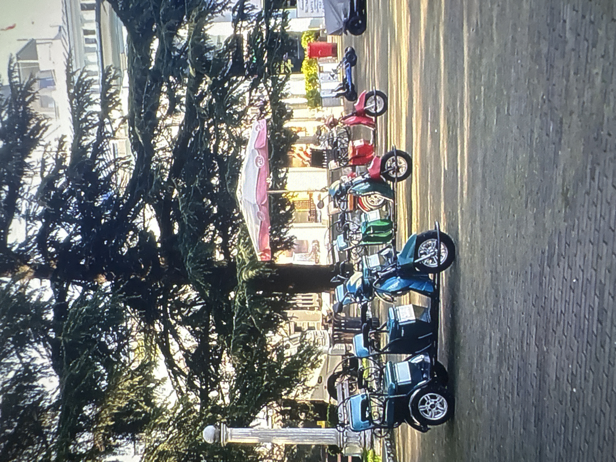The image depicts an outdoor scene, likely in the city of Batumi, Georgia, featuring a row of parked electric scooters or three-wheeled vehicles in the foreground. The vehicles are predominantly green and teal, with some appearing to be designed for two passengers with a single driver. Behind the vehicles, a large, mature tree with dense foliage dominates the middle ground, casting dappled shadows on the paved ground. In the background, a white building with multiple windows and a white railing is visible. A pink and white umbrella shades a small area further back, and several red cylindrical objects are present. The lighting suggests it is daytime, with sunlight filtering through the tree's branches, creating a warm, slightly hazy atmosphere. No people are directly visible in the frame.