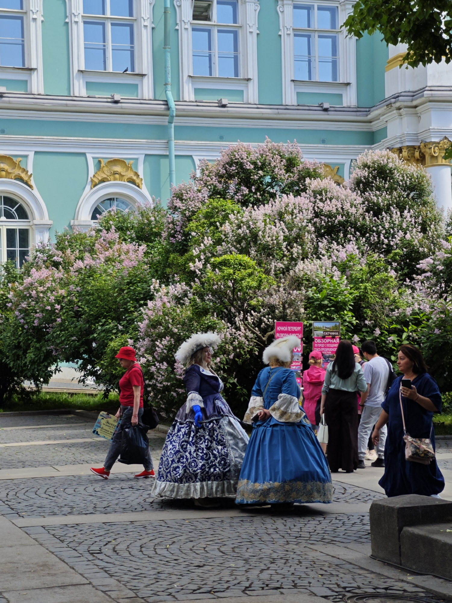 The image depicts a lively scene in front of the Catherine Palace (Ekaterininsky Dvorets) in Pushkin, Saint Petersburg, Russia. The palace's distinctive turquoise facade with white classical detailing and ornate gold elements serves as a grand backdrop. In the foreground, several individuals are present. Two women are dressed in elaborate 18th-century style gowns, one in blue with intricate silver embroidery and a large white feathered headdress, and another in a patterned blue and white dress. A woman in a red hat and red shirt walks past them, carrying a shopping bag and a black plastic bag. Behind them, a group of people in modern attire, including men in white shirts and women in casual clothing, are visible, some looking at informational signs posted near a lush green bush covered in pink blossoms. The ground is paved with cobblestones. The overall atmosphere suggests a tourist attraction or a special event related to historical reenactment.