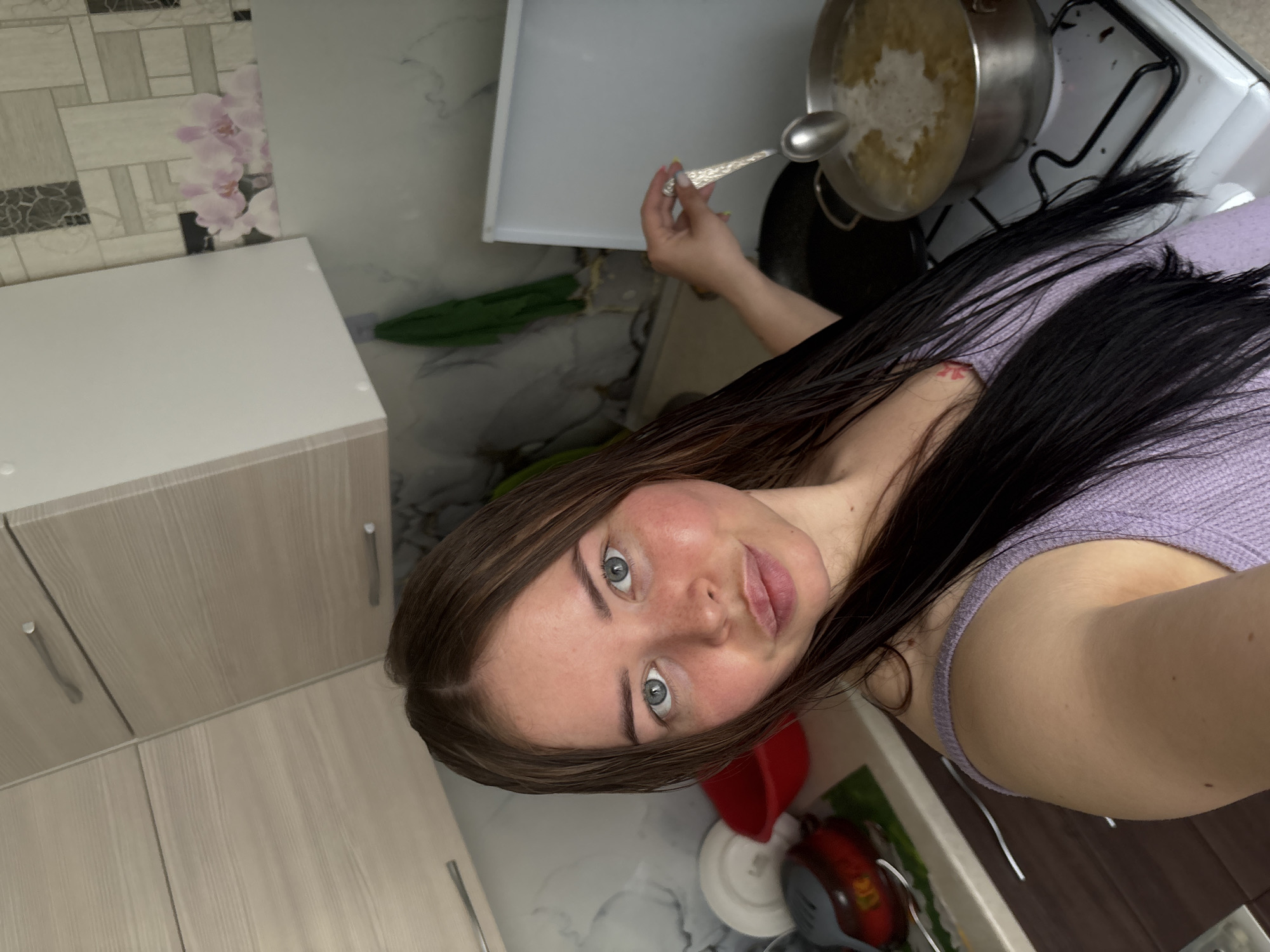A woman with brown hair and blue eyes is in a kitchen. She is holding a spoon and appears to be cooking something in a pot on a stove. Kitchen cabinets are visible in the background, and there is wallpaper with a floral design. The kitchen appears to be located in Izhevsk, Russia.