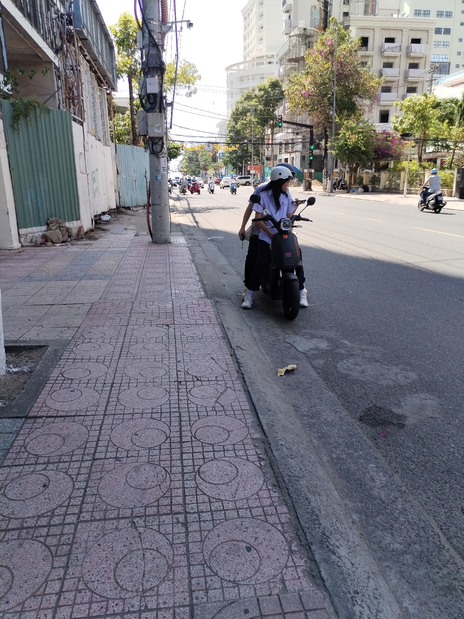The photograph depicts a street scene in what appears to be a ward of Nha Trang City, Vietnam. A young person wearing a white helmet, blue and white collared shirt, and dark trousers stands on the street, straddling a black electric moped. They appear to be looking at a mobile phone attached to the moped.
The scene includes a sidewalk paved with patterned tiles, a curb separating the sidewalk from the asphalt street, and a utility pole with attached electrical boxes. Other motorbikes are visible further down the road, along with buildings in the background. The sky is clear and the light is bright. A piece of trash lies on the street near the curb.