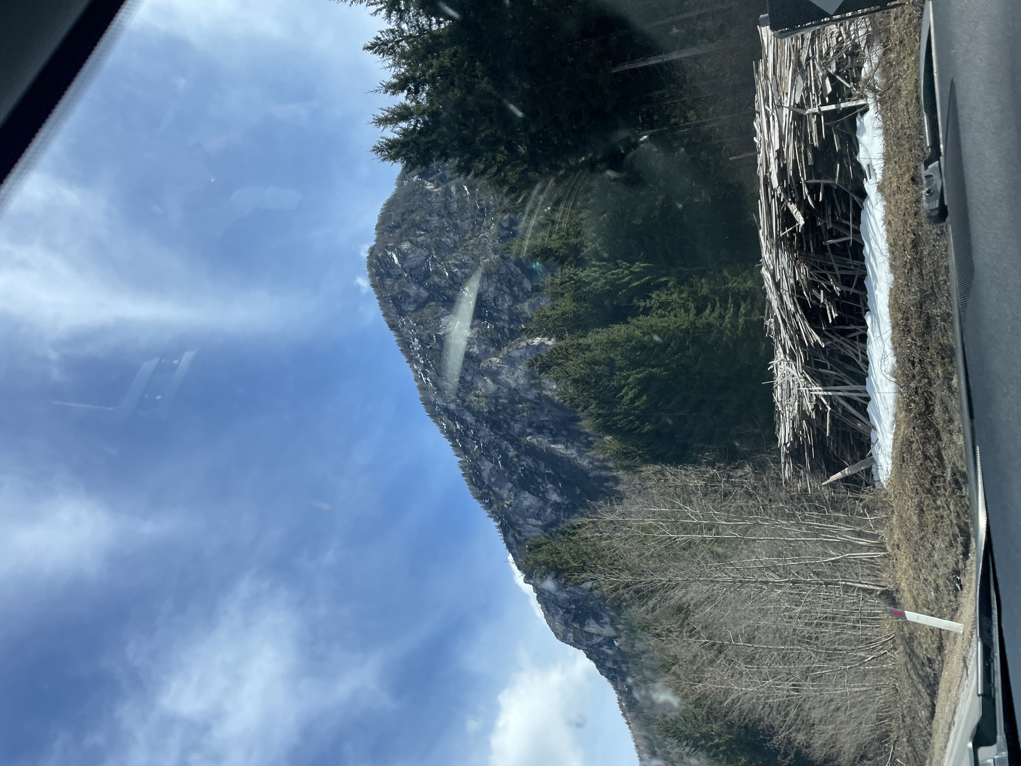 The image shows a mountain partially covered in trees and some remaining snow, captured from inside a vehicle, likely a car, as part of the car's window frame is visible. The sky is blue with scattered clouds. Beside the road, there is a structure made of wood and branches, partially covered in snow. A road sign is also visible. It is likely that this is in the area of Santo Stefano di Cadore, Italy, given the presence of mountainous terrain and trees. The scene is a rural or mountainous area, depicting a natural landscape viewed from inside a car.