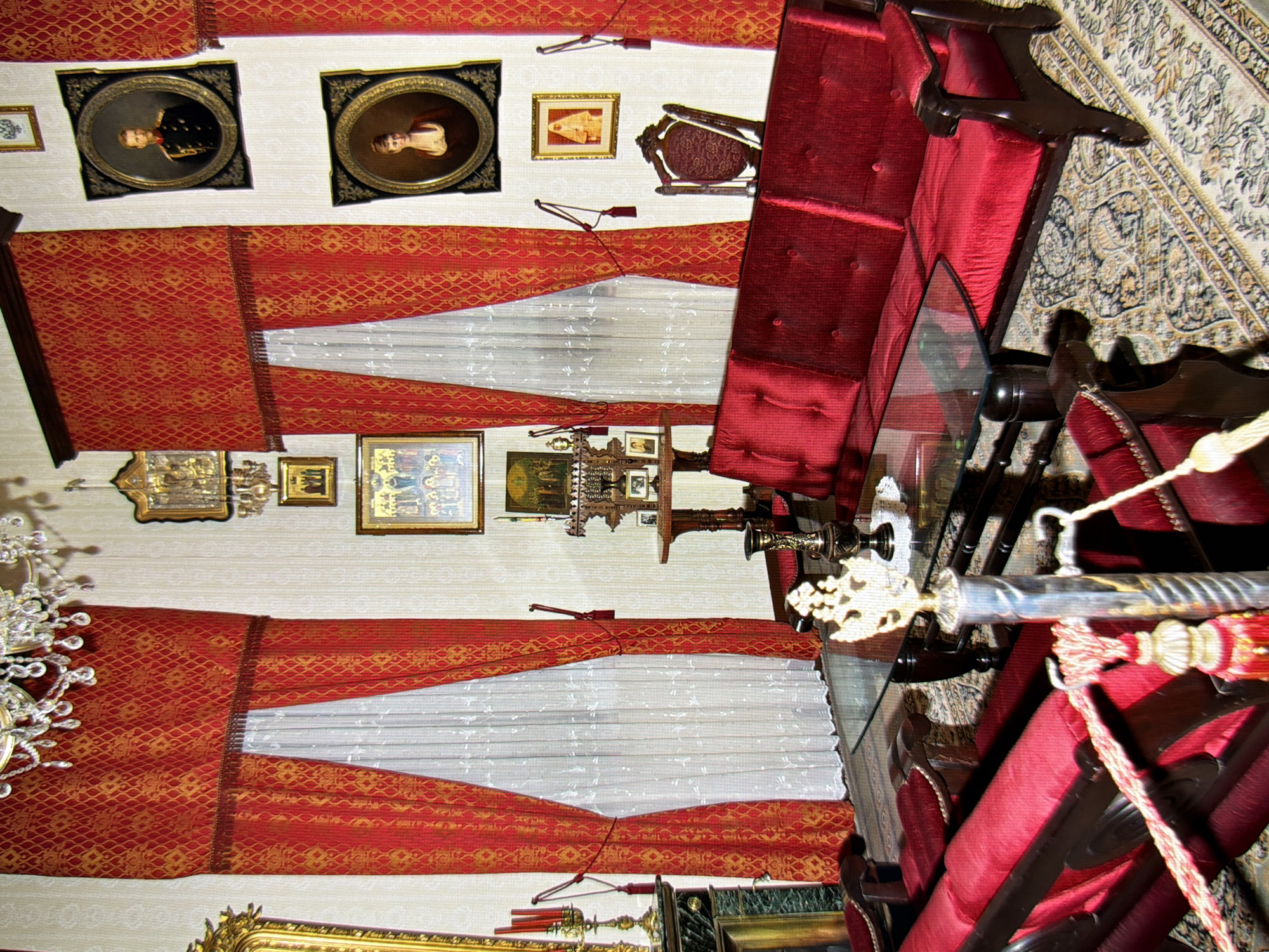 The image depicts a luxurious interior, likely a formal living room or salon, adorned with rich red velvet draperies and ornate furnishings. The room is decorated with several framed portraits and religious icons displayed on the walls. In the center of the room, a large, plush red sofa and a matching chaise lounge are arranged around a glass-topped coffee table. A decorative crystal chandelier hangs from the ceiling, and an elaborate gilded mirror is visible on the left side of the frame. The floor is covered with an intricately patterned rug. The overall impression is one of opulence and historical grandeur. This scene is located in the Mariyinsky Palace, the official ceremonial residence of the President of Ukraine, situated in Kyiv, Ukraine. There are no individuals visible in the photograph.