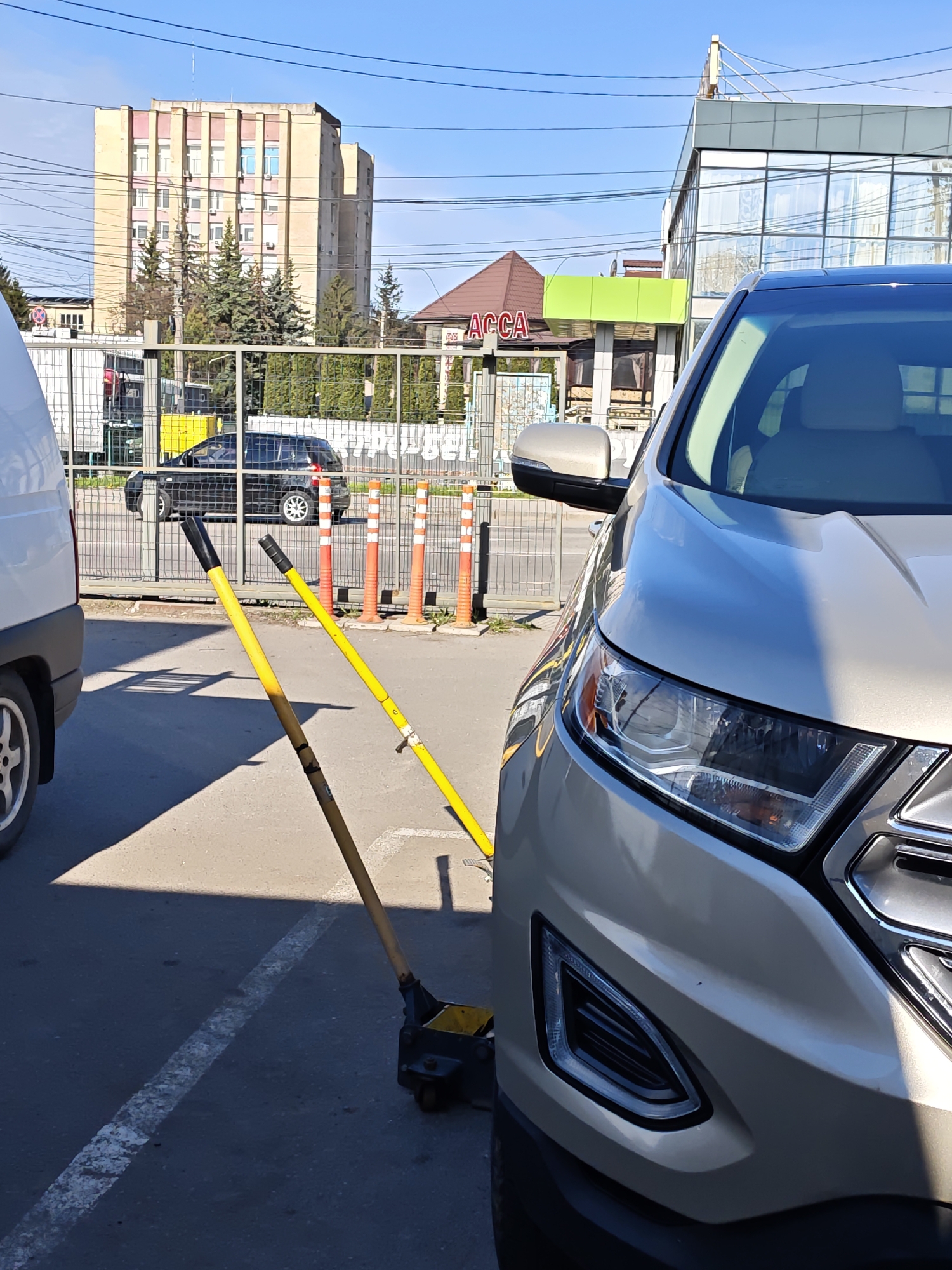 The image shows a parking lot scene in what appears to be Vinnytsia, Ukraine, based on the "ACCA" signage. A light-colored Ford Edge is parked, occupying the right side of the frame. Adjacent to it on the left is a white van, partially visible. A black hydraulic car jack with two yellow handles is positioned near the Ford's front end, seemingly placed between parking spaces. Behind the vehicles, an iron fence with orange traffic cones is visible. Through the fence, a black SUV and a multi-story building with a beige facade and pink trim are discernible. The sky is clear and blue, indicating a sunny day. The architecture in the background suggests an urban environment. The image focuses on the vehicles and parking lot elements with no people visible.