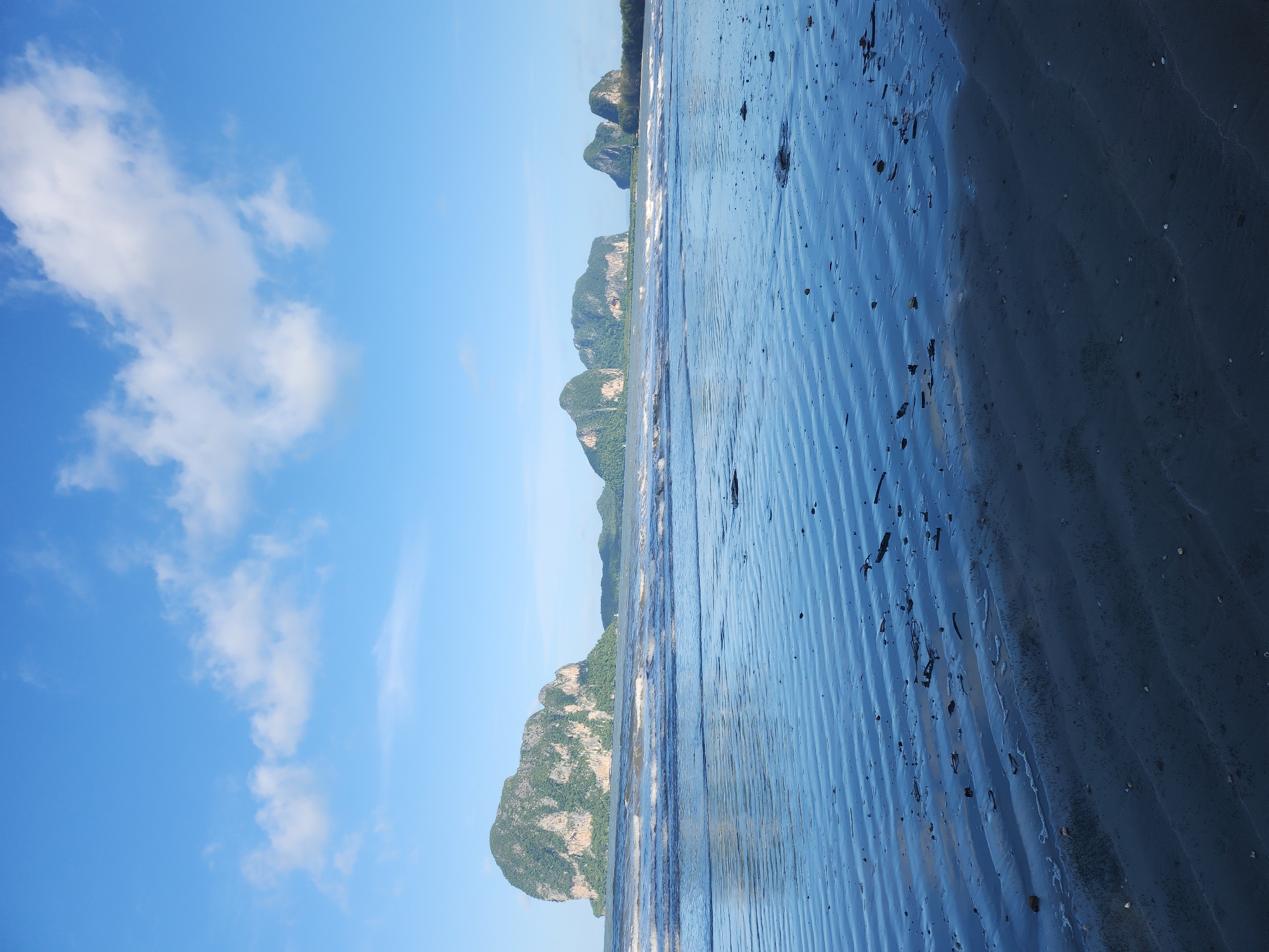 The image depicts a serene beach scene at Hat Pak Meng, Thailand, under a clear blue sky dotted with fluffy white clouds. In the foreground, the wet sand exhibits distinct ripple patterns left by receding tides, with scattered debris like small pieces of wood and shells visible. The sea in the midground is calm, with gentle waves lapping at the shore, reflecting the sky's blue hue. In the distance, a series of lush, green, rocky islands rise dramatically from the ocean. The largest island, prominent on the left, is a significant geological feature. The overall atmosphere is tranquil, capturing the natural beauty of the coastline. No people are present in the scene.