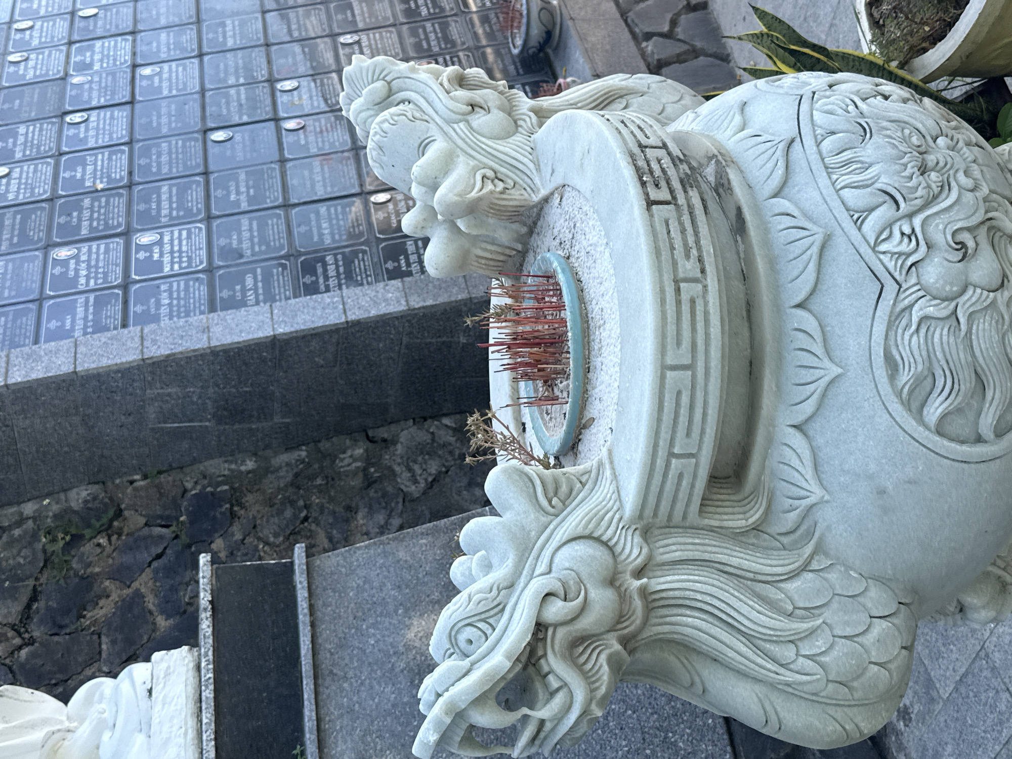 The image shows a stone incense burner, intricately carved in the shape of a dragon, with red incense sticks burning in it. The incense burner is made of light-colored stone, possibly marble, and features detailed carvings of dragon heads on either side, along with scales and decorative patterns. In the background, a wall of memorial plaques is visible, suggesting a cemetery or a memorial site. The plaques are arranged in rows and appear to have names and dates inscribed on them. The overall setting seems to be outdoors, with a stone wall and some greenery in the foreground. The presence of the memorial plaques strongly suggests this scene is located in a cemetery in the city of Nha Trang Ward, Vietnam.