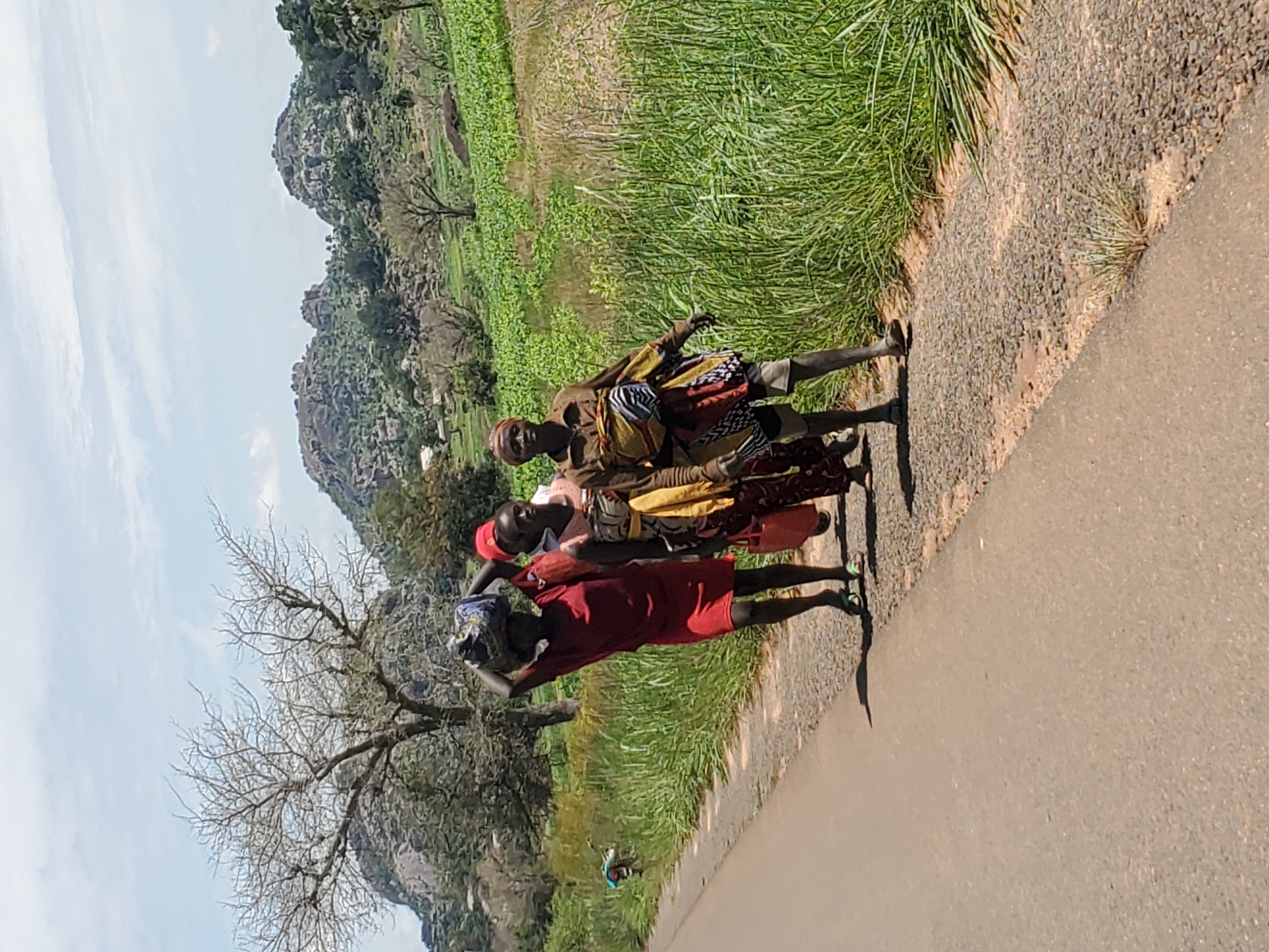 The image depicts four women walking along a paved road, carrying bundles on their heads. The road is bordered by a gravel shoulder and lush green vegetation. In the background, a hilly landscape with rocky outcrops and scattered trees is visible, along with agricultural fields. The sky is partly cloudy. The overall scene suggests a rural or semi-rural environment, possibly in the vicinity of Pankshin, Nigeria, given the geographical context. The women are dressed in colorful clothing, and their posture indicates they are engaged in travel or transport.