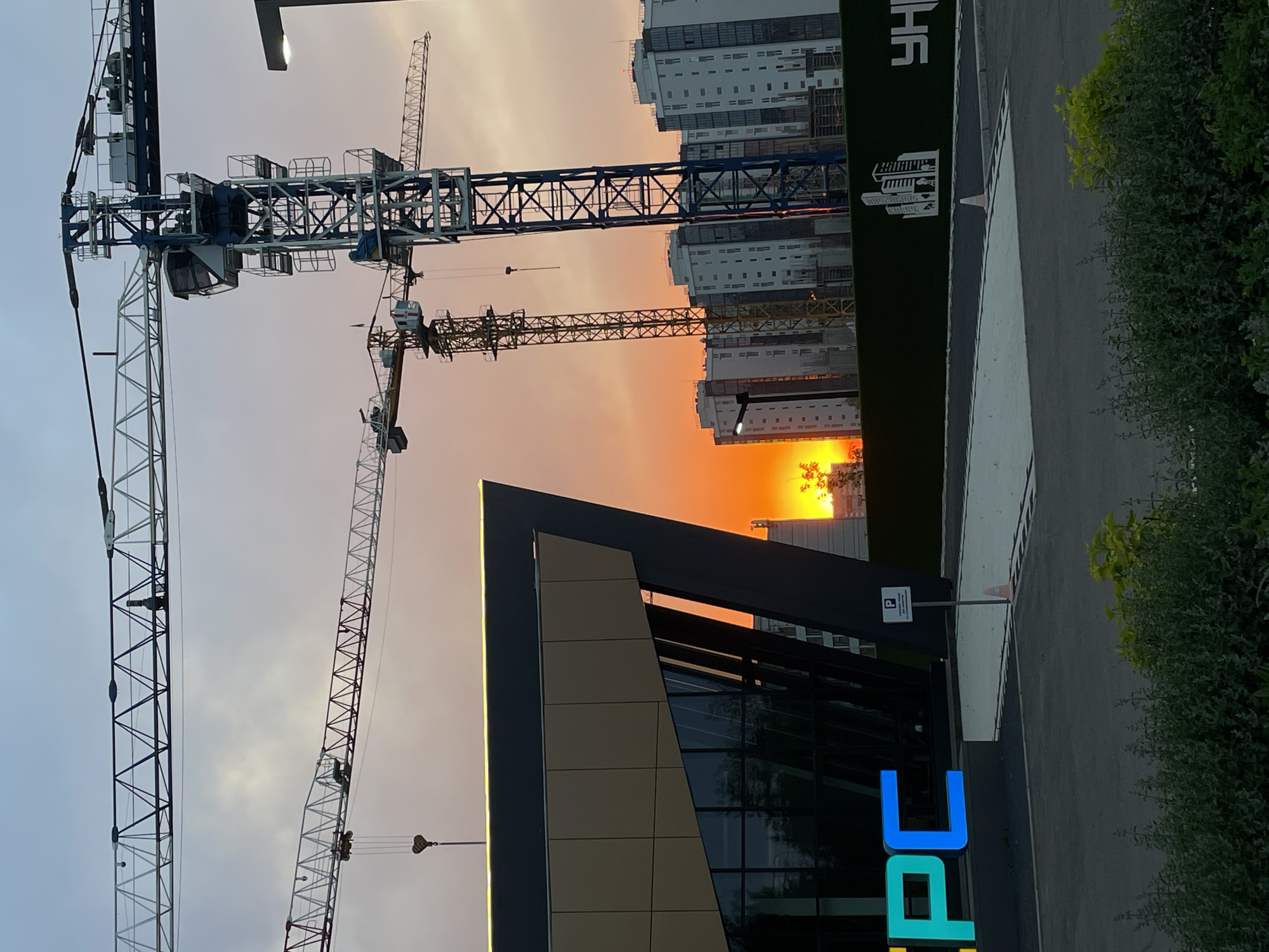 The image depicts a construction site in the city of Krasnoyarsk, Russia, at sunset. In the foreground, a modern building with a dark facade and large glass windows is visible on the left. The building has illuminated signage spelling "CPC" in large, colorful letters. To the right of the building, a parking sign is mounted on a pole.
Dominating the midground and background are several large construction cranes, silhouetted against a vibrant orange sunset. These cranes are part of the ongoing construction of residential buildings, which are visible in the distance as tall, white apartment blocks. A green fence runs along the edge of the construction site, featuring a white graphic of buildings and Cyrillic lettering.
The sky is a gradient of soft blues and oranges, indicating either dusk or dawn. The overall scene captures the dynamic activity of urban development against a natural, atmospheric backdrop. There are no people visible in the image. The ground in the foreground is paved, and there is some low-lying vegetation. The lighting suggests it is late afternoon or early evening.
