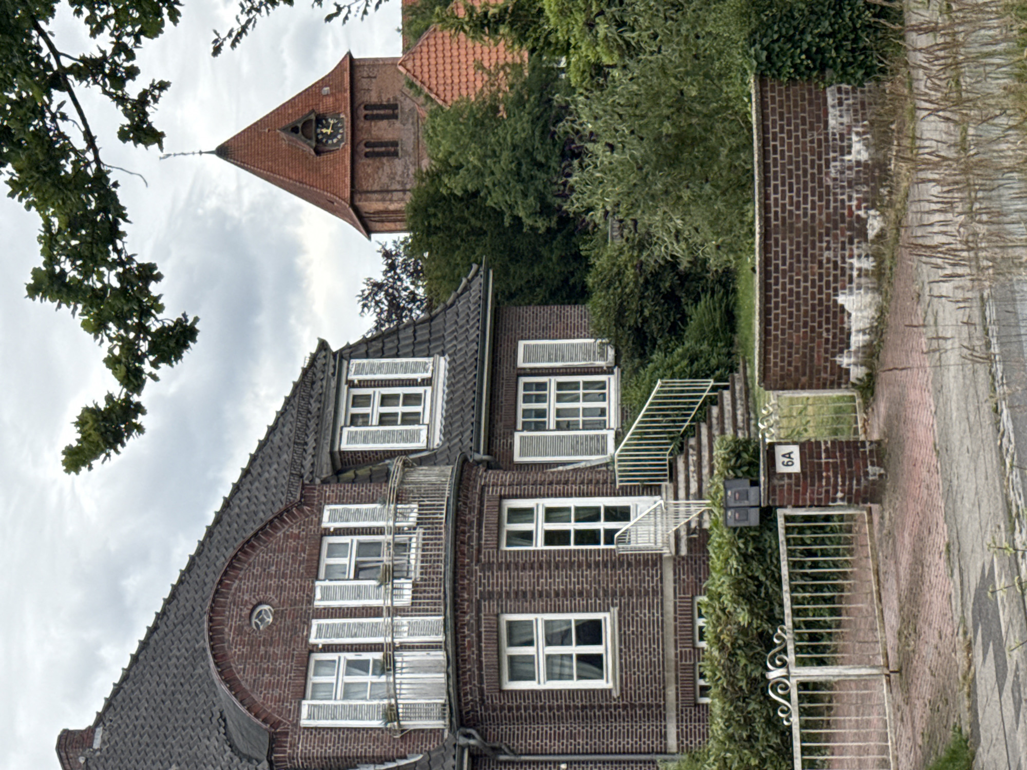 The image shows a large, ornate brick house with white-shuttered windows and a curved balcony. A steep set of stairs leads to the front door, with mailboxes at the base of the stairs. In the background, a red-tiled church steeple rises above lush green trees, suggesting a serene, possibly rural or suburban setting. The overall impression is one of a well-maintained, historic property. The house is located at 6A in Bassum, Germany.