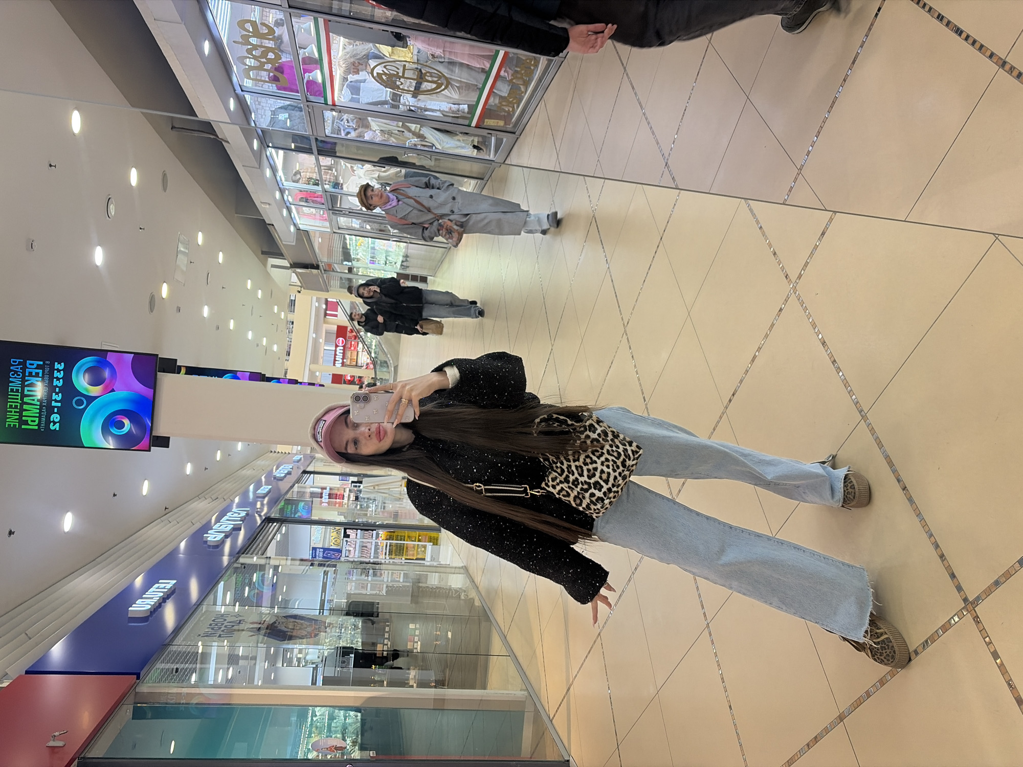 This media file captures a scene within a brightly lit shopping mall, likely in Saint Petersburg, Russia. The primary subject is a young woman taking a selfie in a large, reflective wall. The image is taken by another person, evidenced by their feet (wearing dark trousers and sneakers) visible at the bottom right and part of their arm at the top left.
The young woman, with long dark hair, is wearing a dark, possibly sparkly, long-sleeved top, light blue wide-leg jeans, and leopard-print platform shoes or sneakers. She sports a light pink baseball cap and carries a leopard-print shoulder bag. She holds a light-colored smartphone (possibly pink/purple) up to her face, angling it to capture her reflection, with a neutral to slightly pouting expression.
The large mirror reflects the mall environment, including other shoppers and various store fronts. In the background reflection, another woman in a grey coat and hat is visible, along with two other indistinct figures further away. The tiled floor features a diamond pattern with decorative mosaic strips. Overhead, numerous recessed spotlights illuminate the space.
Visible text in the image includes:
- On a large digital display to the left, Cyrillic text such as "В ОБЛАСТИ МЕДИЦИНЫ" (In the field of medicine), "БУЗИЕТЕНИЕ" (Buzzing), and "МУИМ," along with an alphanumeric sequence "222-31-es."
- A reflected shop sign at the top right clearly shows "PELOSO."
- Another reflected shop sign further back partially reads "Auss."
- A blue shop sign, also reflected, displays white Cyrillic text, appearing to be "УЛИЯНА" (Ulyana) and possibly "УЛИЧНАЯ МОДА" (Street Fashion).
The activity centers around the young woman posing for a photograph while simultaneously taking a selfie in the mall's reflective surface, amidst the usual foot traffic of a commercial center.
