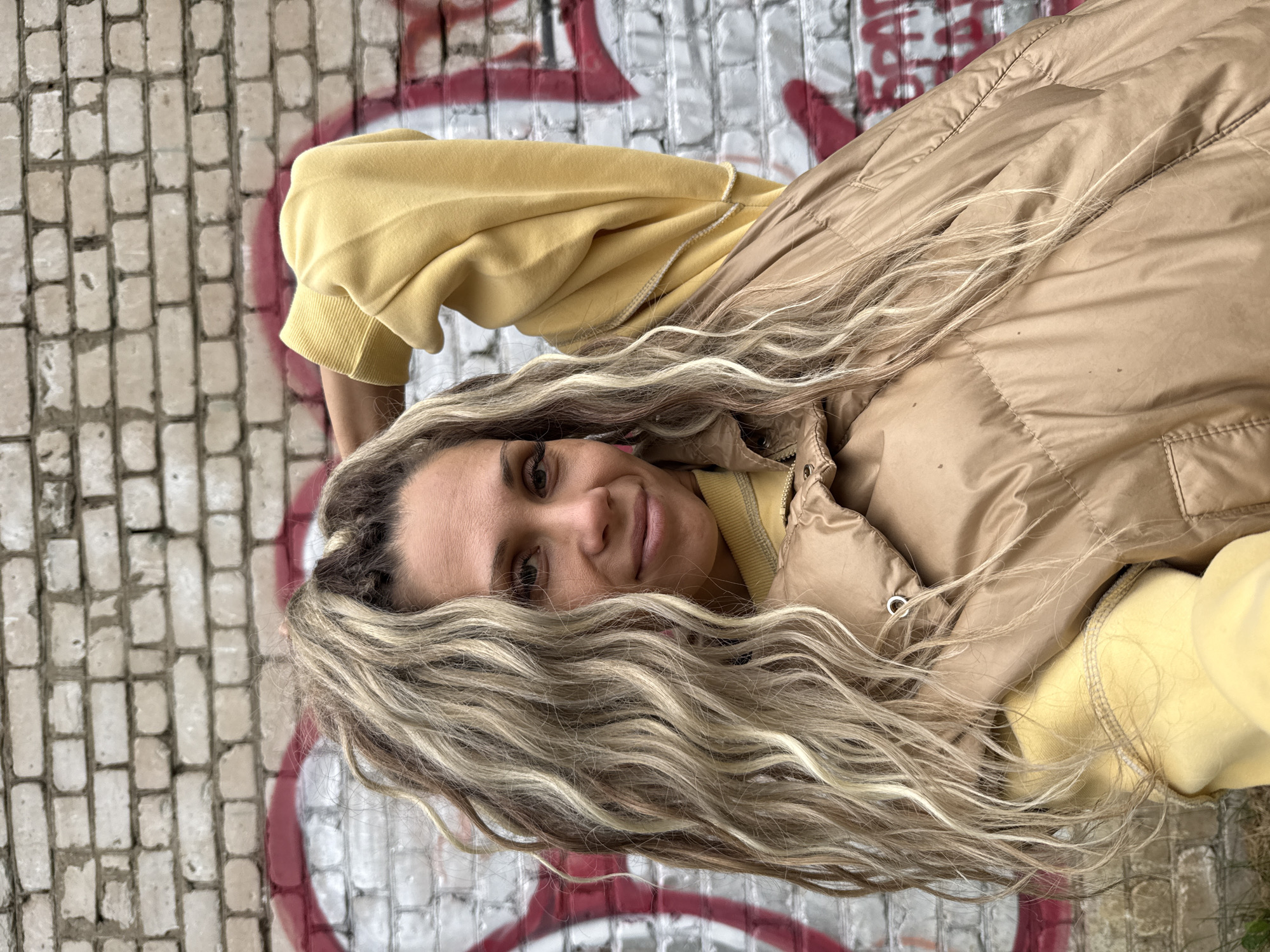 A woman with blonde, wavy hair stands in front of a graffiti-covered brick wall. She is wearing a yellow sweatshirt and a tan puffer vest. Her right arm is raised behind her head. The wall is made of light-colored bricks and has red graffiti sprayed on it. The scene appears to be outdoors in an urban environment.