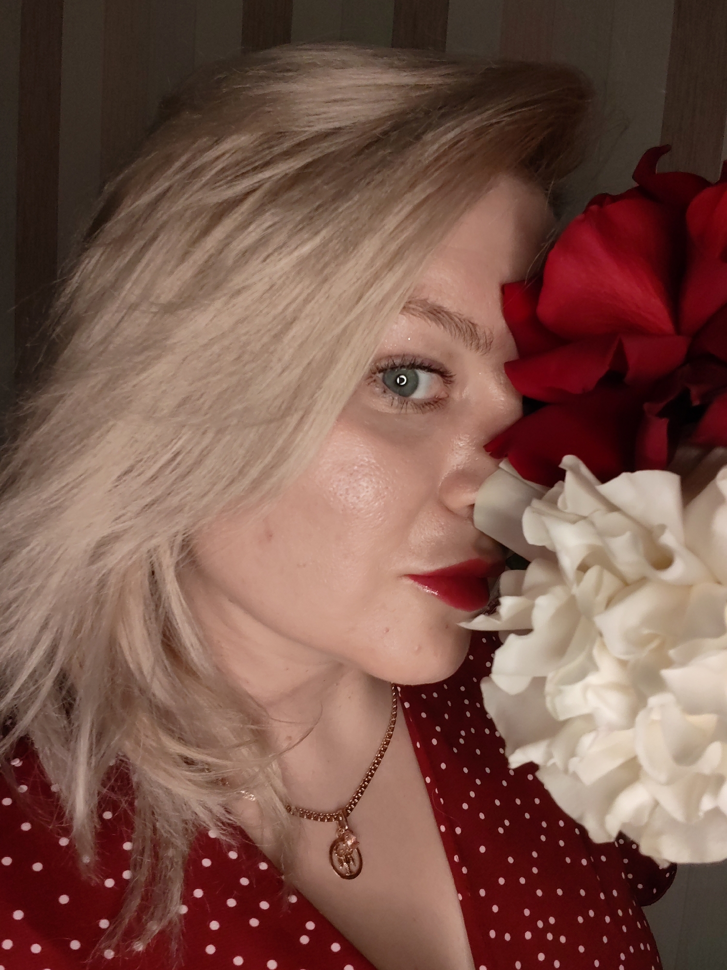 A close-up portrait features a young woman with fair skin, voluminous blonde hair, and striking light blue-green eyes that gaze directly at the viewer. Her face is partially framed and subtly obscured by two large, lush flowers: a deep red one positioned to the right of her face, and a creamy white one below it, both appearing to be either roses or peonies. She is wearing bright red lipstick and her natural skin texture is visible.
The woman is dressed in a red garment with white polka dots, and a delicate gold-colored chain necklace with a small, circular pendant featuring a raised, intricate motif, possibly an animal figure, adorns her neck. The setting appears to be indoors, with a dimly lit background showcasing vertical dark and light stripes, suggestive of wallpaper or blinds. The foreground, including the woman and the flowers, is well-lit, indicating an artificial light source. The woman is posing for the camera, creating a stylized and intimate composition. The image is contextualized to Izhevsk, Russia. There is no visible text in the image.