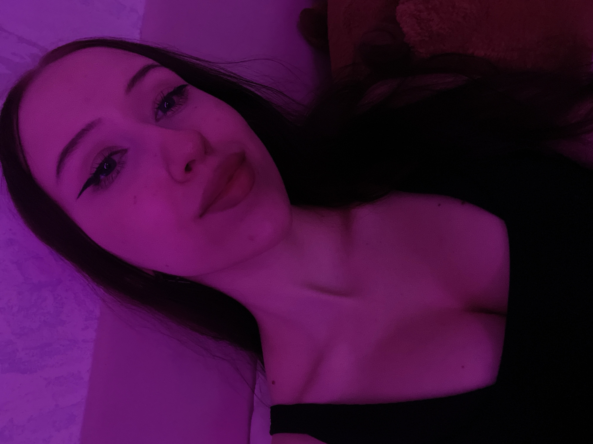 A young woman with long, dark hair, wearing a black top, is pictured in a close-up shot. She has winged eyeliner and her lips are slightly parted. The scene is illuminated by a vibrant pink light, creating a purplish hue across her skin and the background. The background appears to be a textured wall. There is no discernible connection to Votkinsk, Russia.