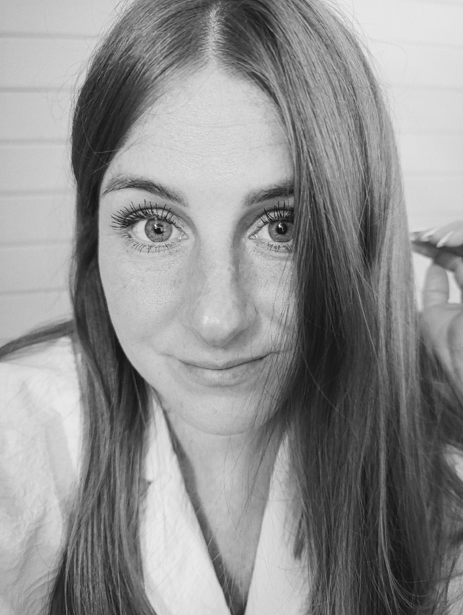 A black and white close-up shot shows the face of a young woman. She has long, straight brown hair that is parted in the middle and falls around her face. She is wearing a white shirt. The woman's eyes are wide open and looking directly at the camera. She has freckles on her cheeks and nose. Her lips are slightly parted, and she appears to be smiling faintly. The background is a plain, light-colored wall with horizontal lines. Her left hand is visible in the upper right corner of the frame, and she is touching her hair. The overall impression is a candid, natural portrait.