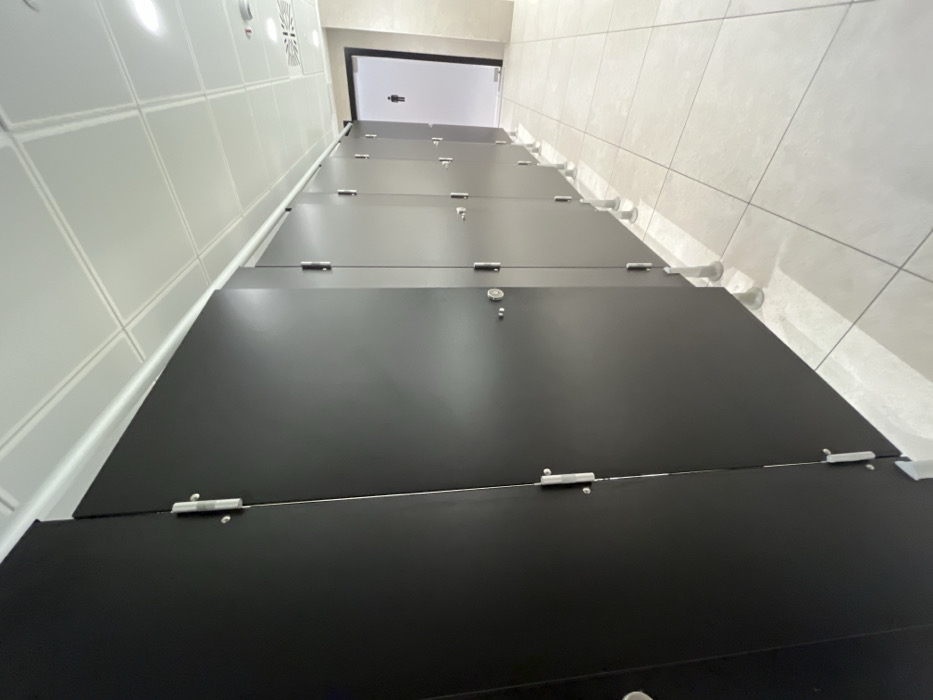 This image depicts the interior of a clean, modern men's public restroom located in Dorno, Italy. The photograph is taken from a low-angle perspective, looking along a lengthy row of black toilet stall doors that recede into the background. Each stall door is equipped with silver-colored hinges and visible latches or locks.
On the left side of the frame, a wall covered in white square tiles extends parallel to the stalls. To the right, a contrasting wall with light beige or grey square tiles is visible, from which white cylindrical supports extend, anchoring the stall partitions. The ceiling is white with a grid pattern, and some light fixtures or vents are discernable.
In the background, a doorway with a clear or frosted glass panel is visible, displaying a prominent male restroom symbol. The restroom appears empty, with no individuals present or any specific activity occurring. The space is brightly lit, and the surfaces of the black stall doors show subtle reflections. The overall impression is one of cleanliness, functionality, and a contemporary design aesthetic.