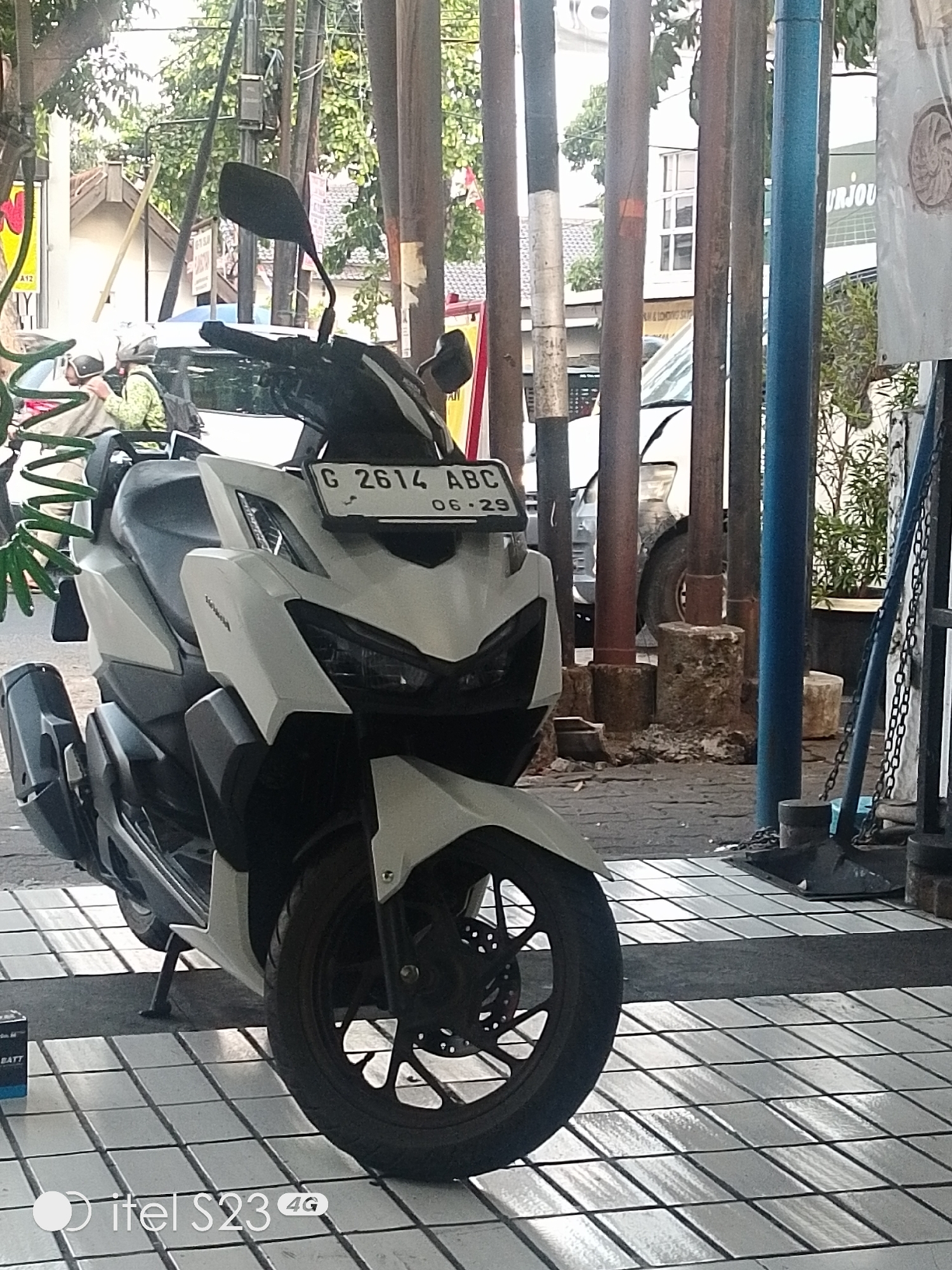 The image shows a white motorcycle parked on a tiled sidewalk in Semarang, Indonesia. The motorcycle has a license plate with the inscription "G 2614 ABC" and "06 - 29". In the background, there are several wooden poles, a white van, and some trees. The scene appears to be on a street, with buildings and a business visible in the distance. The lighting suggests it is daytime, likely in the morning or afternoon.