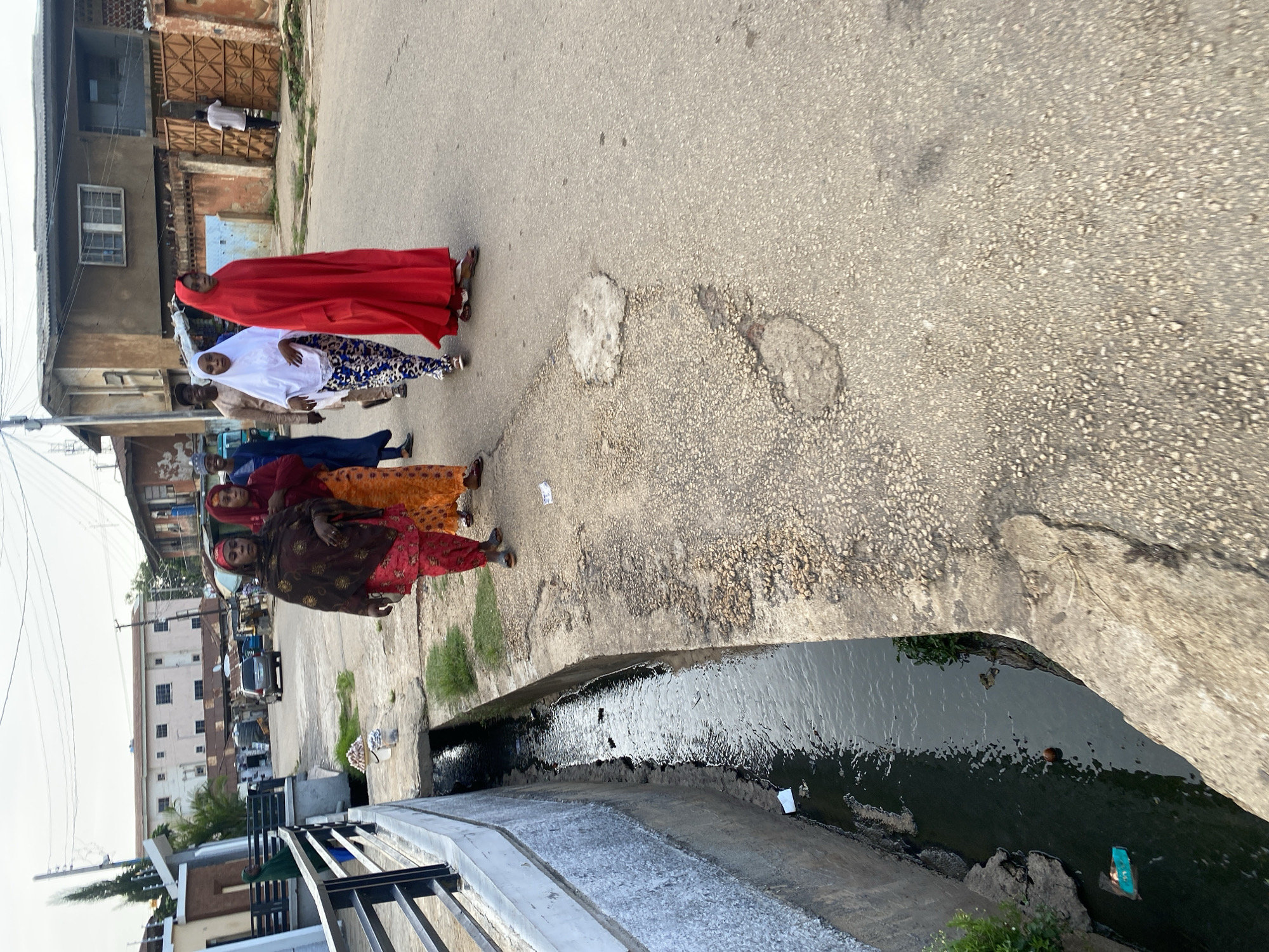 In an urban environment in Jos, Nigeria, a group of women dressed in traditional Islamic attire walk along a paved road. The road shows signs of wear with several potholes. To the left of the road, a concrete drainage channel runs alongside, containing murky water and some debris. In the background, buildings of varying architectural styles and states of repair line the street. Power lines are visible overhead. A few vehicles, including a car and a tricycle, are parked or moving in the distance. The sky is overcast, suggesting a diffused light. The scene depicts everyday life in a Nigerian city, highlighting both its community aspects and the challenges of its infrastructure.