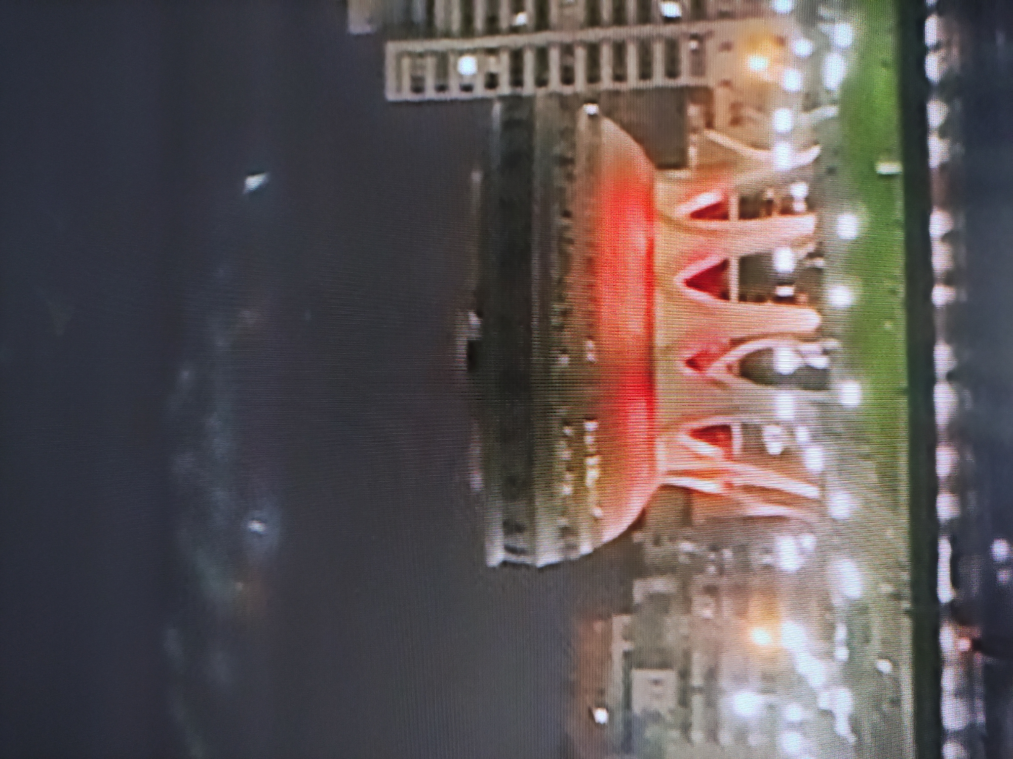 The image depicts an aerial night view of the Ertis Promenade in Pavlodar, Kazakhstan. The most prominent feature is the "Vaza" sculpture, illuminated in red and orange. Adjacent to the sculpture is a green strip of grass and a pathway lined with white lights. In the upper left corner, a portion of a bridge or pier with rectangular lights is visible. The background consists of the dark Irtysh River with some light reflections, and the city with scattered lights.