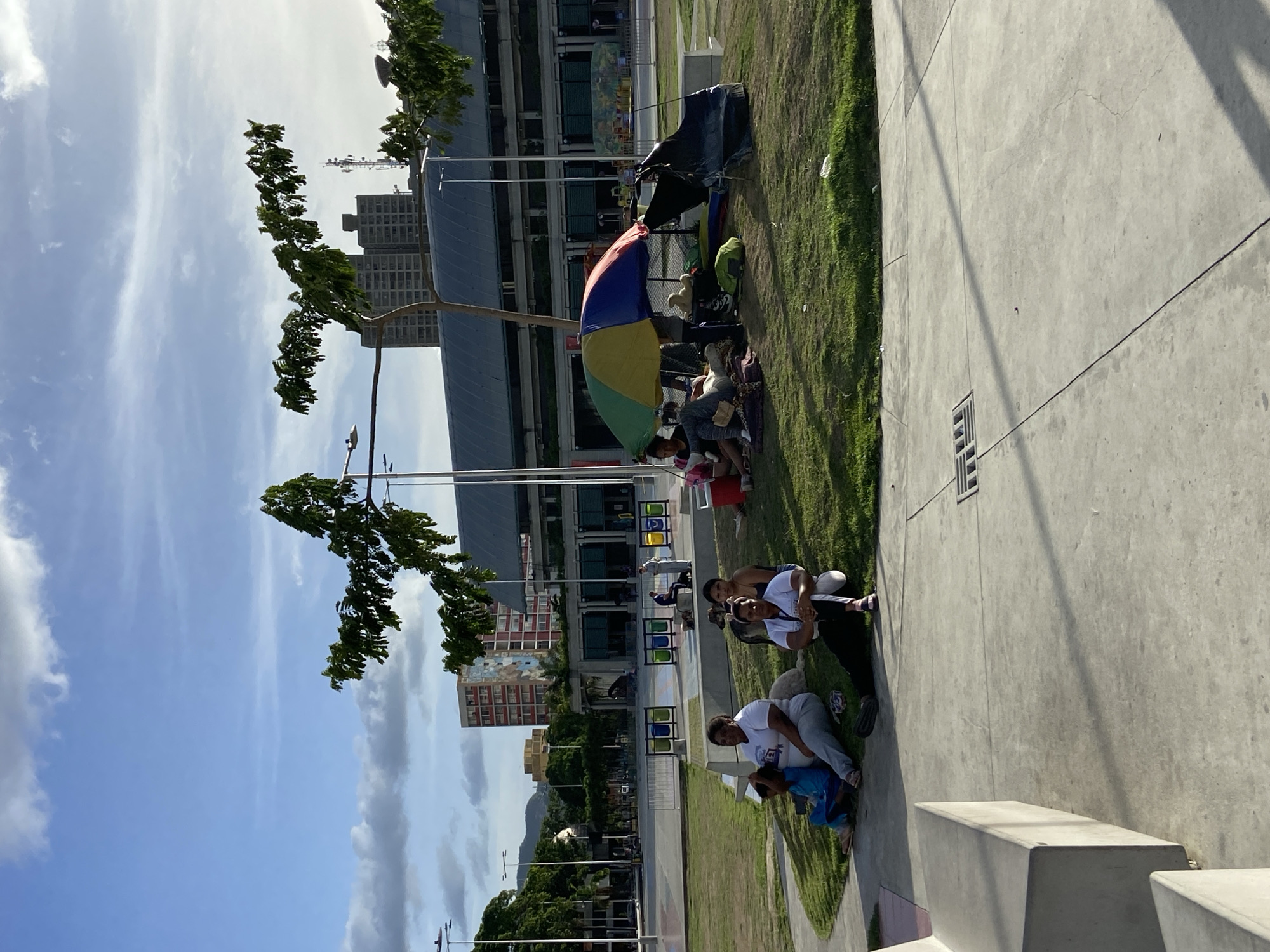 The image depicts a sunny daytime scene in Caracas, Venezuela, with a clear blue sky and scattered clouds. The focus is on a group of people gathered on a grassy area next to a paved walkway. Some individuals are sitting on the grass, while others are partially visible on the walkway. A multi-colored umbrella provides shade for a few people who are seated on the ground, some appearing to be reading or resting.
In the background, modern buildings and trees are visible, suggesting an urban park or public space. A tent-like structure is also present, along with backpacks and other items that might indicate an overnight stay or a gathering. Several people are seen in different poses, some interacting with each other, while others appear more solitary. One group in the foreground on the walkway includes individuals sitting and lying down, with a person in a white shirt and grey pants looking towards the camera. Another person, lying on their back, is partially visible.
The lighting suggests it is midday, with shadows cast on the paved surface. The overall atmosphere appears relaxed, with no obvious signs of distress or conflict among the individuals. The presence of buildings and the style of the architecture provide cues to the urban setting of Caracas. No visible text is present in the image.