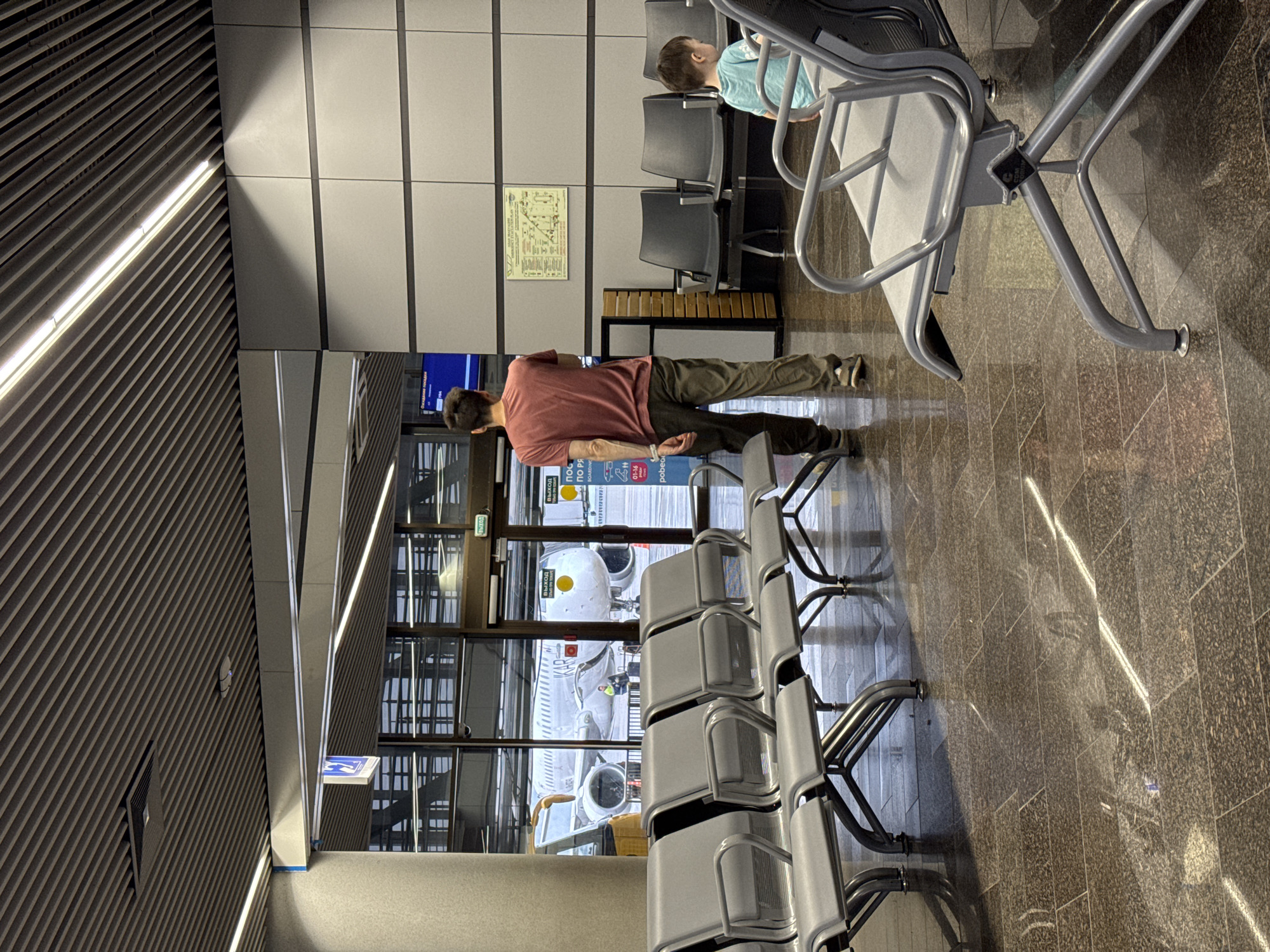 The image captures an airport waiting area, potentially in Sochi, Russia. A man in a reddish-brown shirt and dark pants stands in front of a window. Outside the window, an airplane is visible. A row of empty, gray metal chairs runs along the right side of the image, with the airplane seen through the window behind them. Another person, possibly a child wearing a light blue shirt, is seated in a chair behind the man. Wall panels and a diagram are visible on the wall behind the people. The floor is made of brown marble-like tiles.